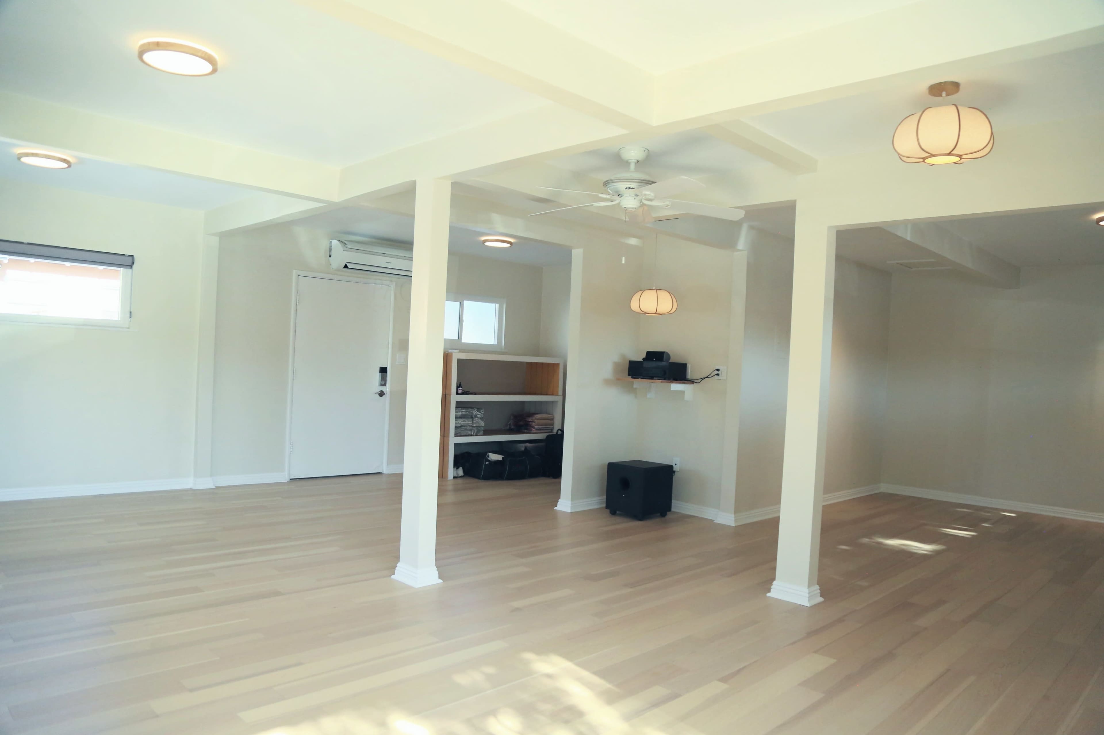 The image shows an empty, well-lit room with wooden flooring, white walls, and a ceiling fan, featuring two pendant lights and a small workspace in the corner.