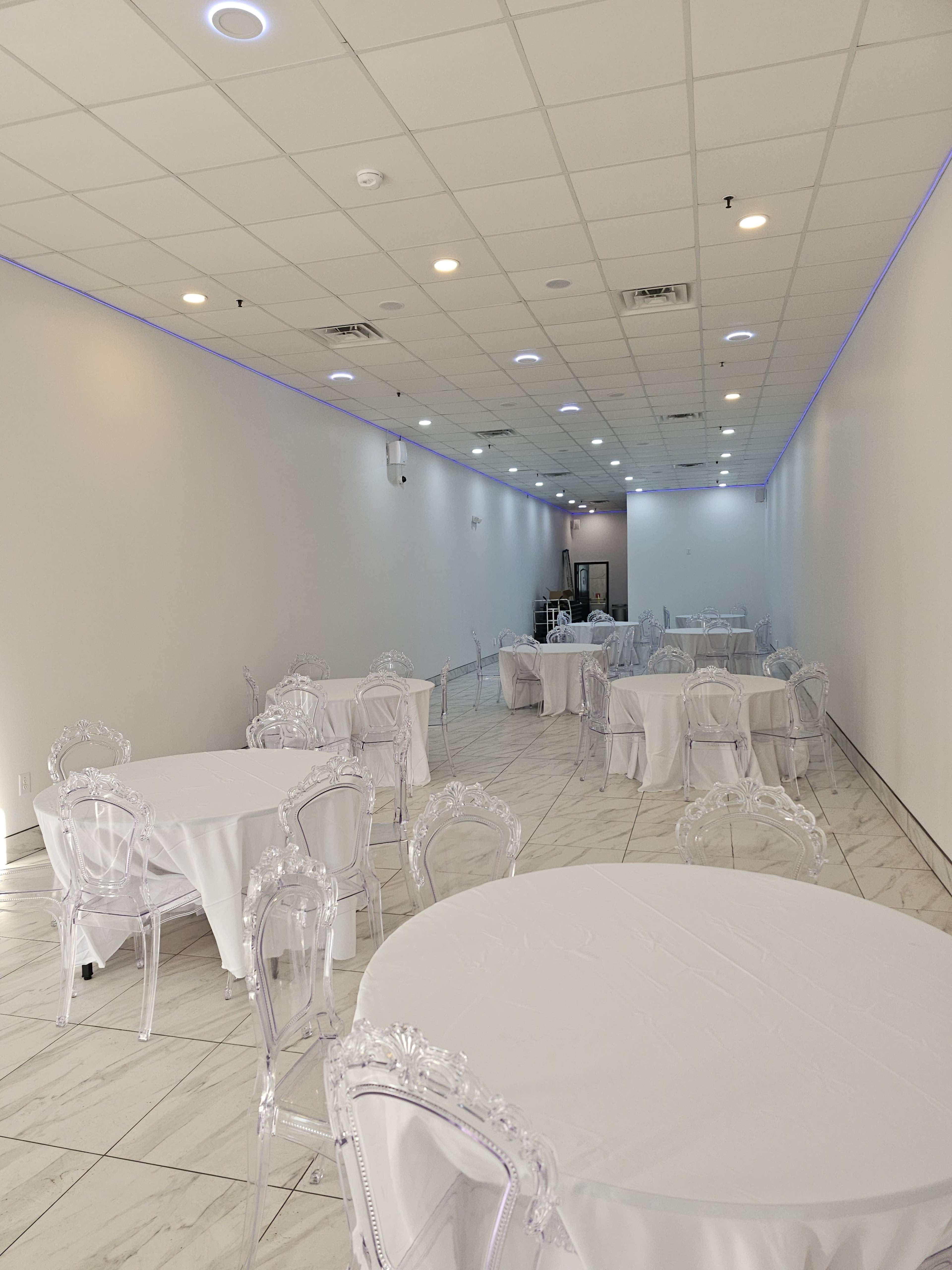 The image shows an empty event space featuring white tables covered with tablecloths and clear plastic chairs arranged throughout the room, with soft lighting along the ceiling.