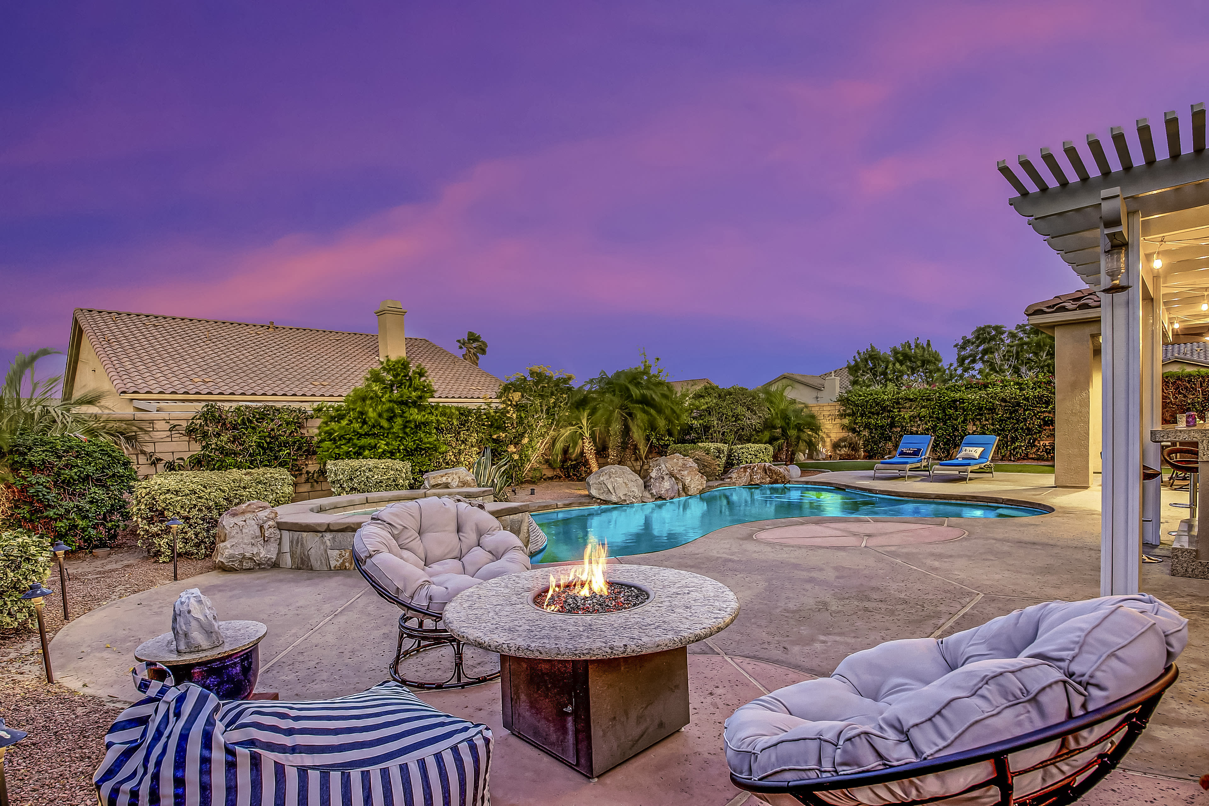 A backyard features a swimming pool, a fire pit surrounded by cushioned chairs, and vibrant sunset colors in the sky.