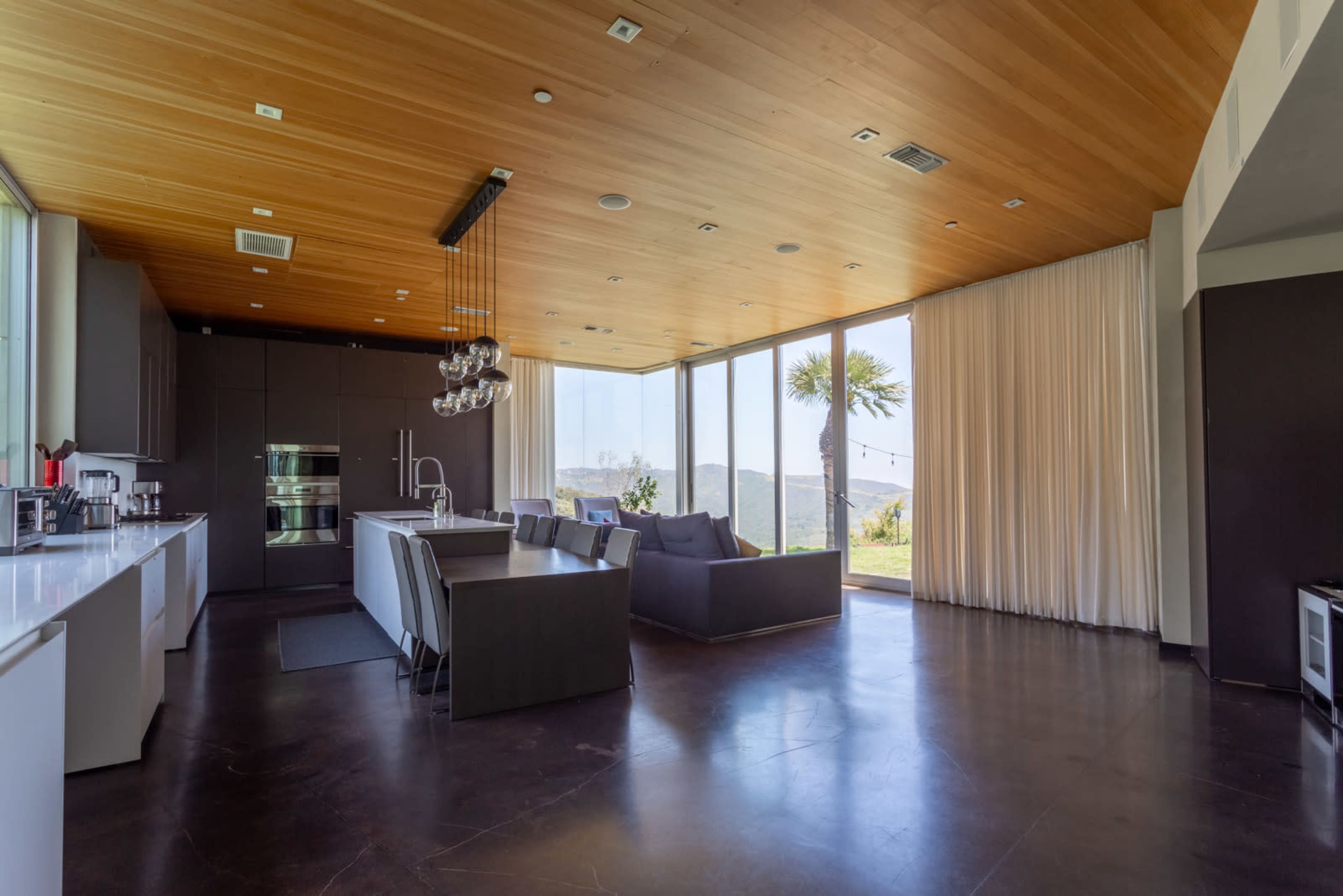 A modern open-concept kitchen and living area with large windows, hardwood ceilings, and contemporary furniture.