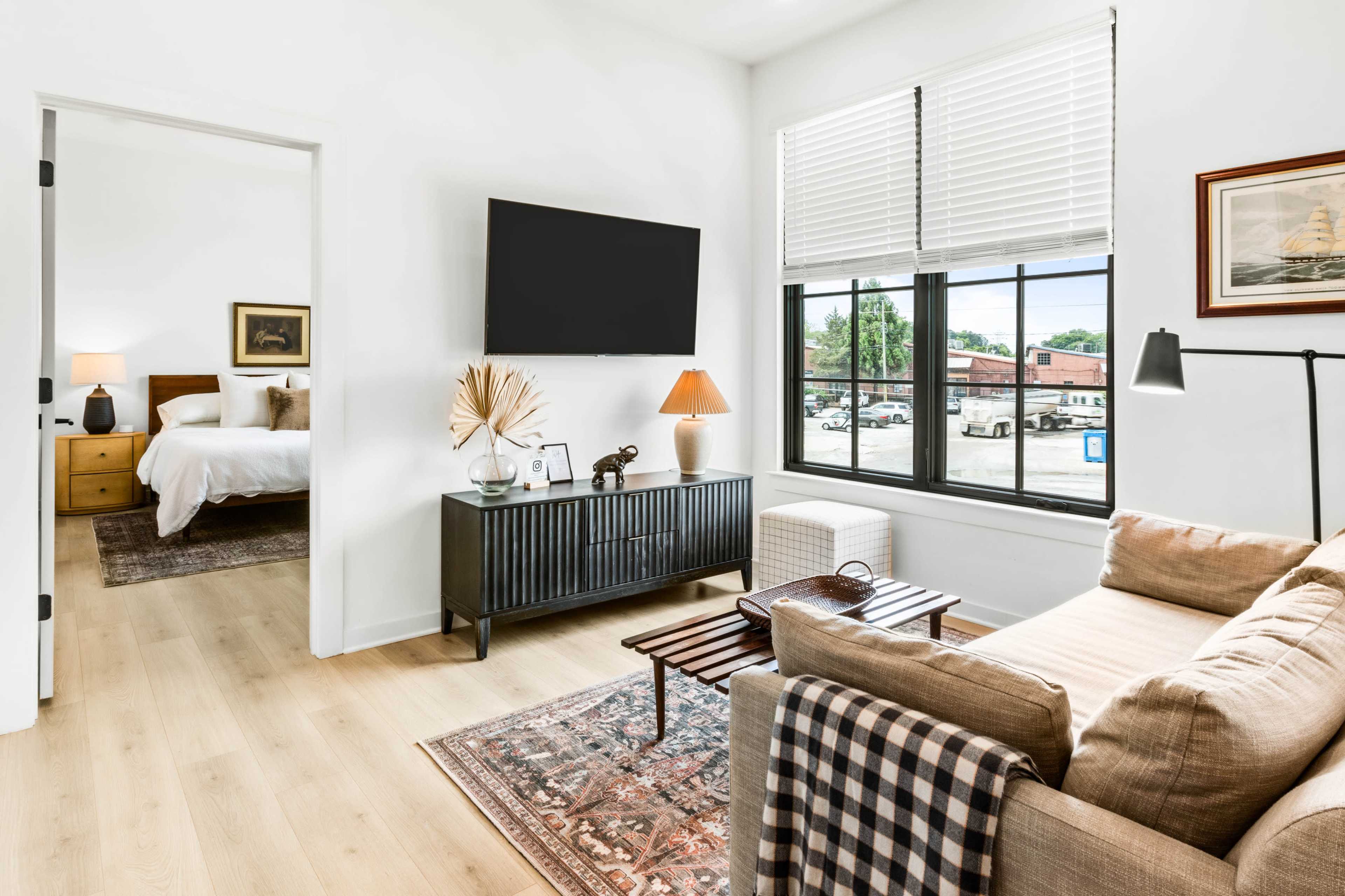 The image shows a modern living room featuring a beige sofa, a coffee table, and a large wall-mounted television, with a doorway leading to a bedroom in the background.