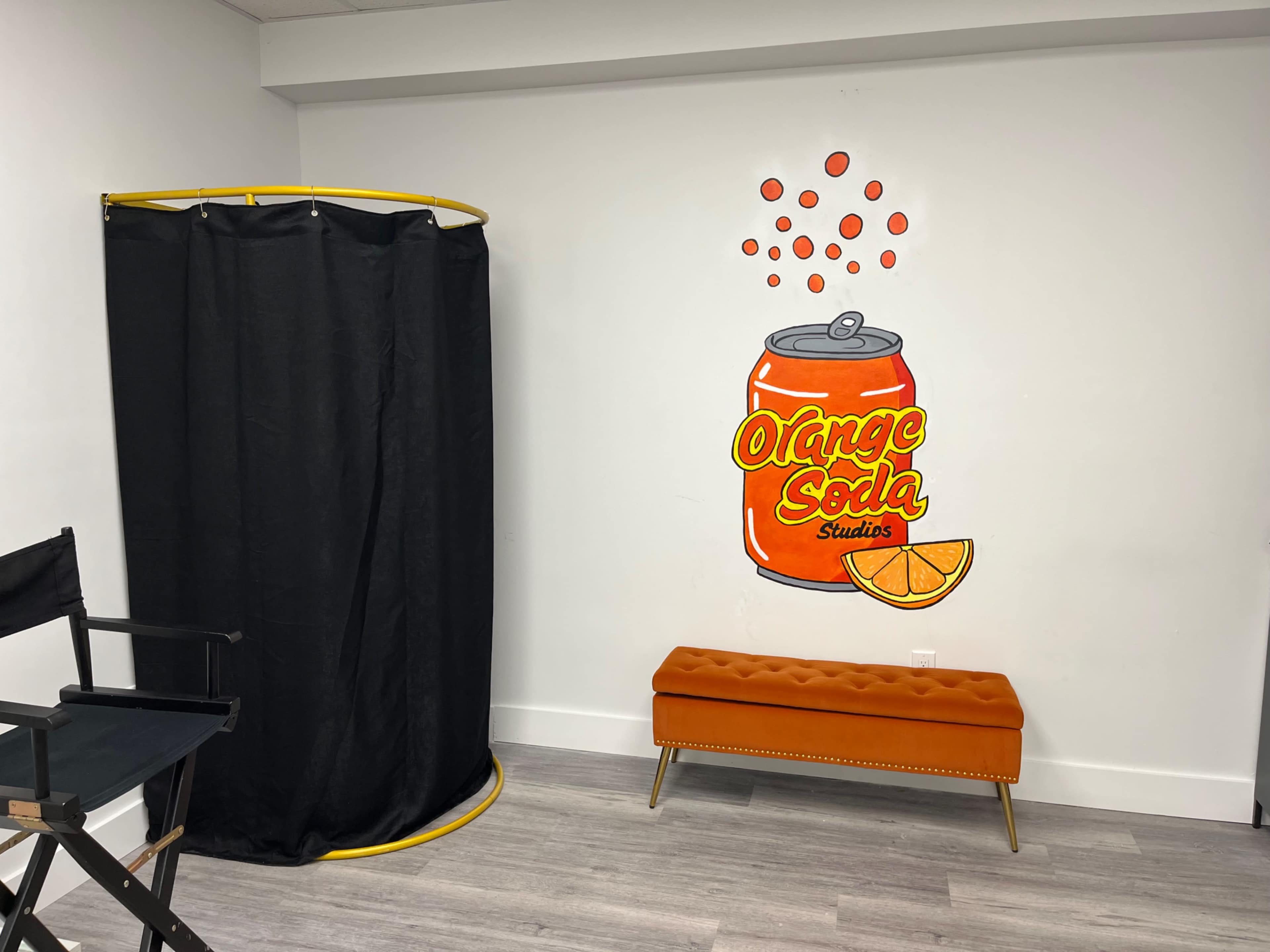 The image shows a small room featuring a black curtain enclosure, an orange tufted bench, and a mural of an orange soda can with bubbles on the wall.