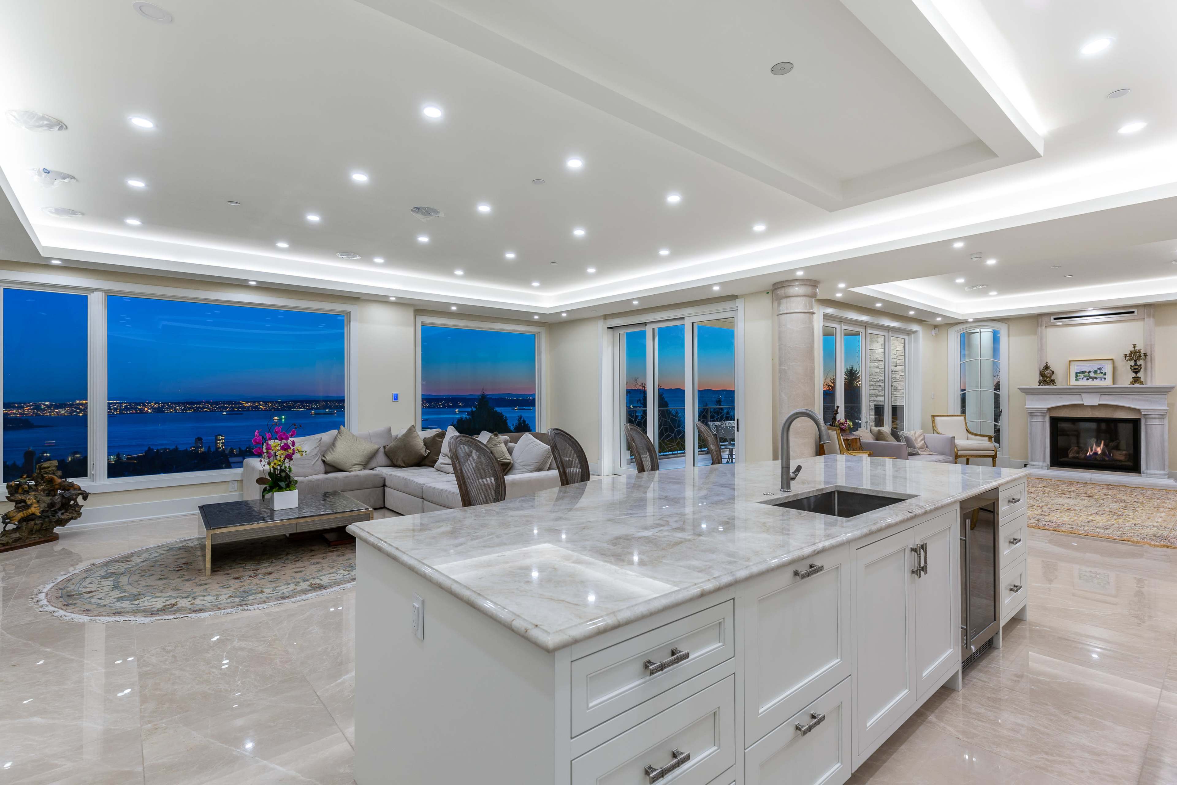 A spacious modern living room features large windows with views of a body of water and city skyline, illuminated by recessed lighting and a fireplace.