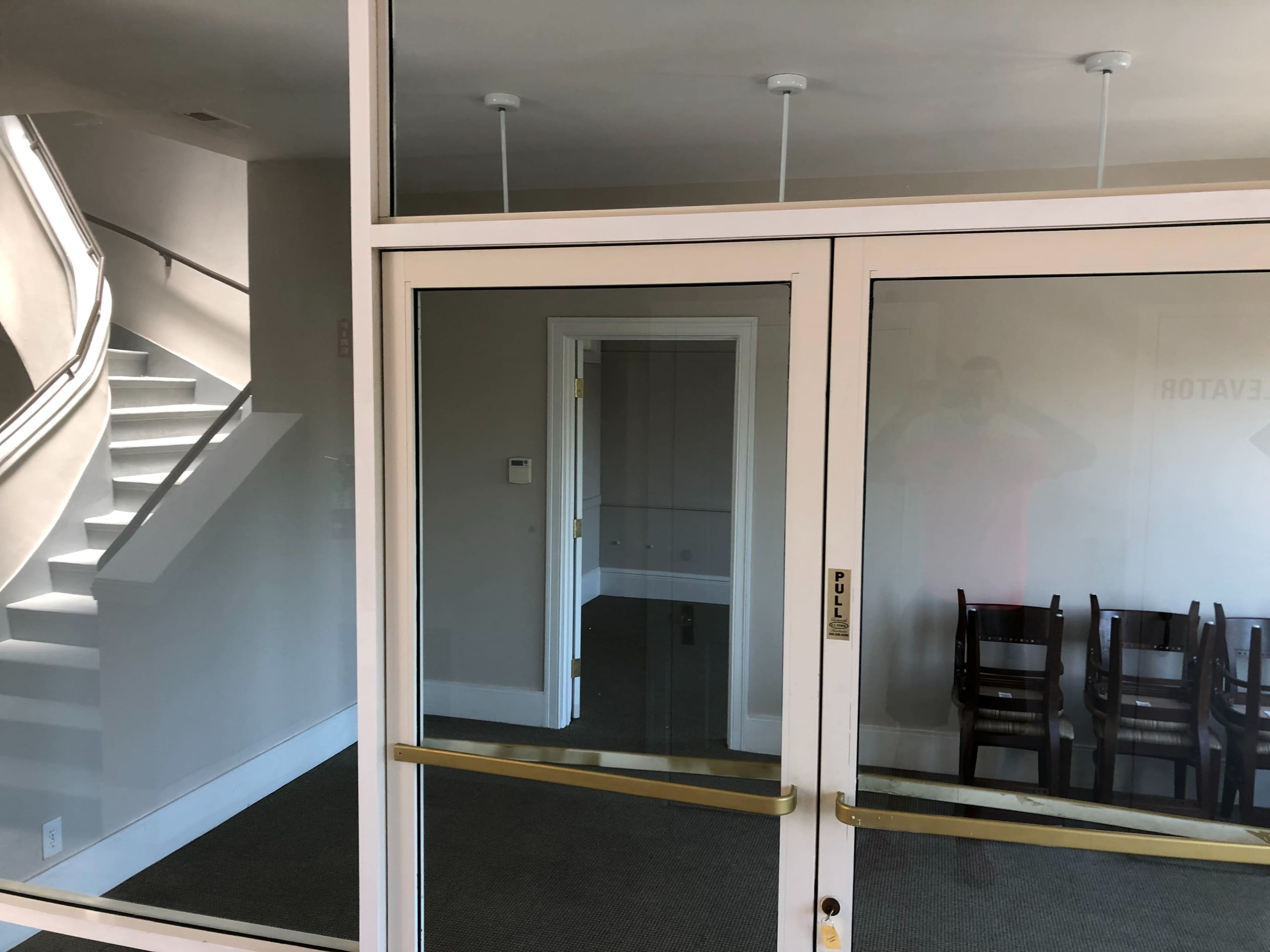 A glass entrance with a reflection of a staircase and a room featuring several stacked chairs behind it.