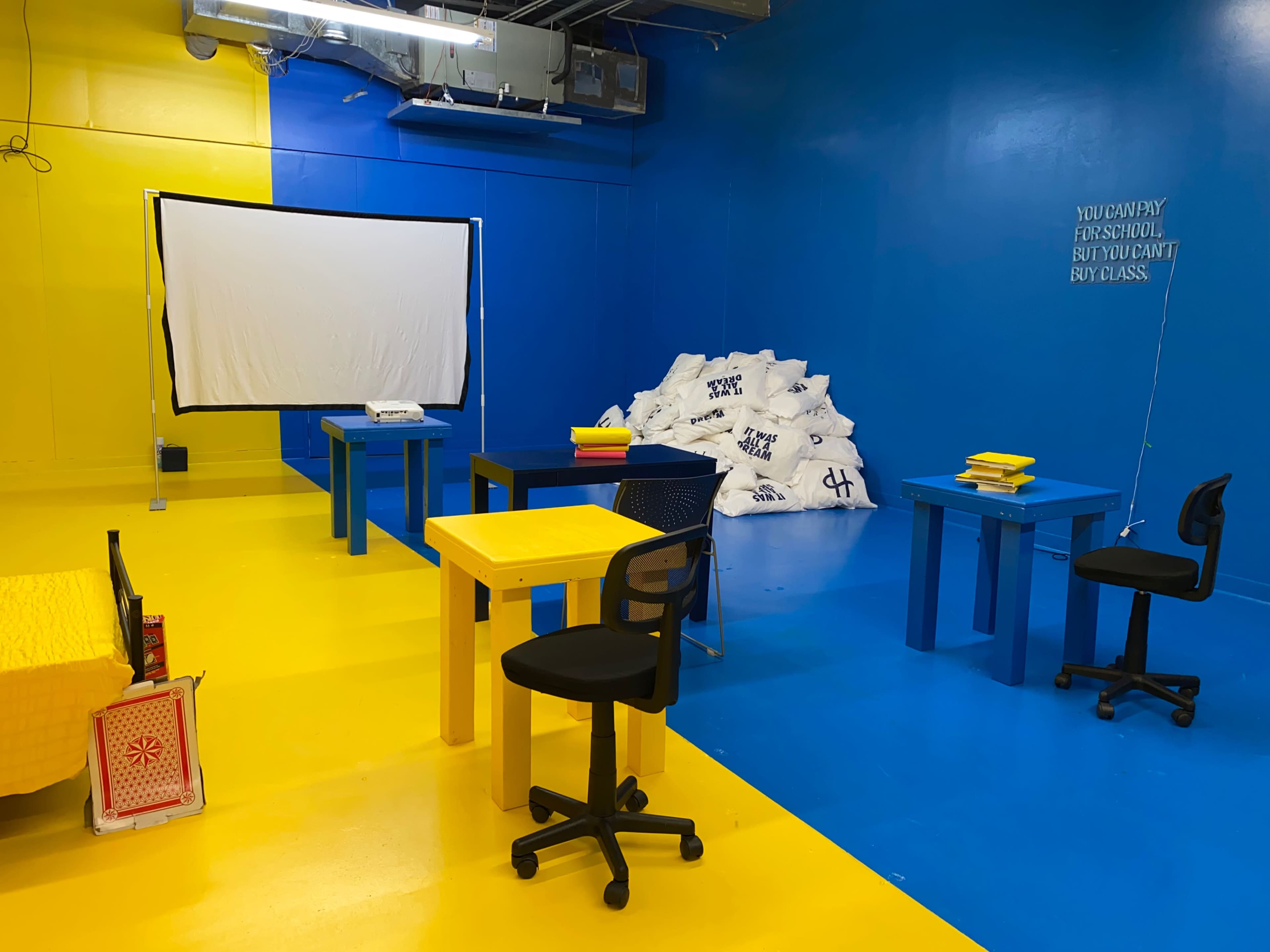 The room features bright yellow and blue walls, a projector screen, several tables in contrasting colors, and a pile of materials in the corner.