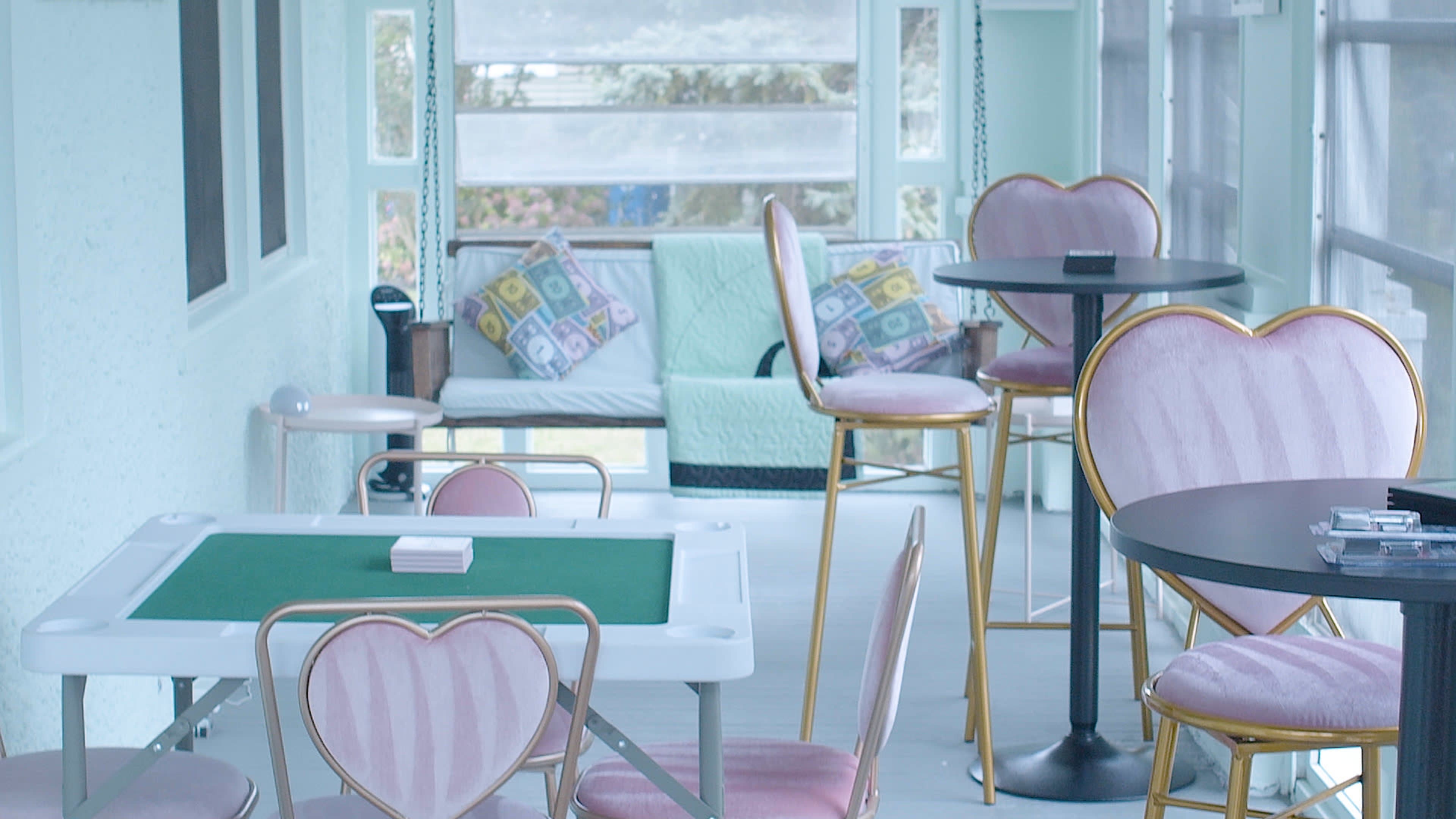 The image shows a light-colored lounge area featuring heart-shaped chairs, a green felt card table, and a cozy seating arrangement with colorful cushions.