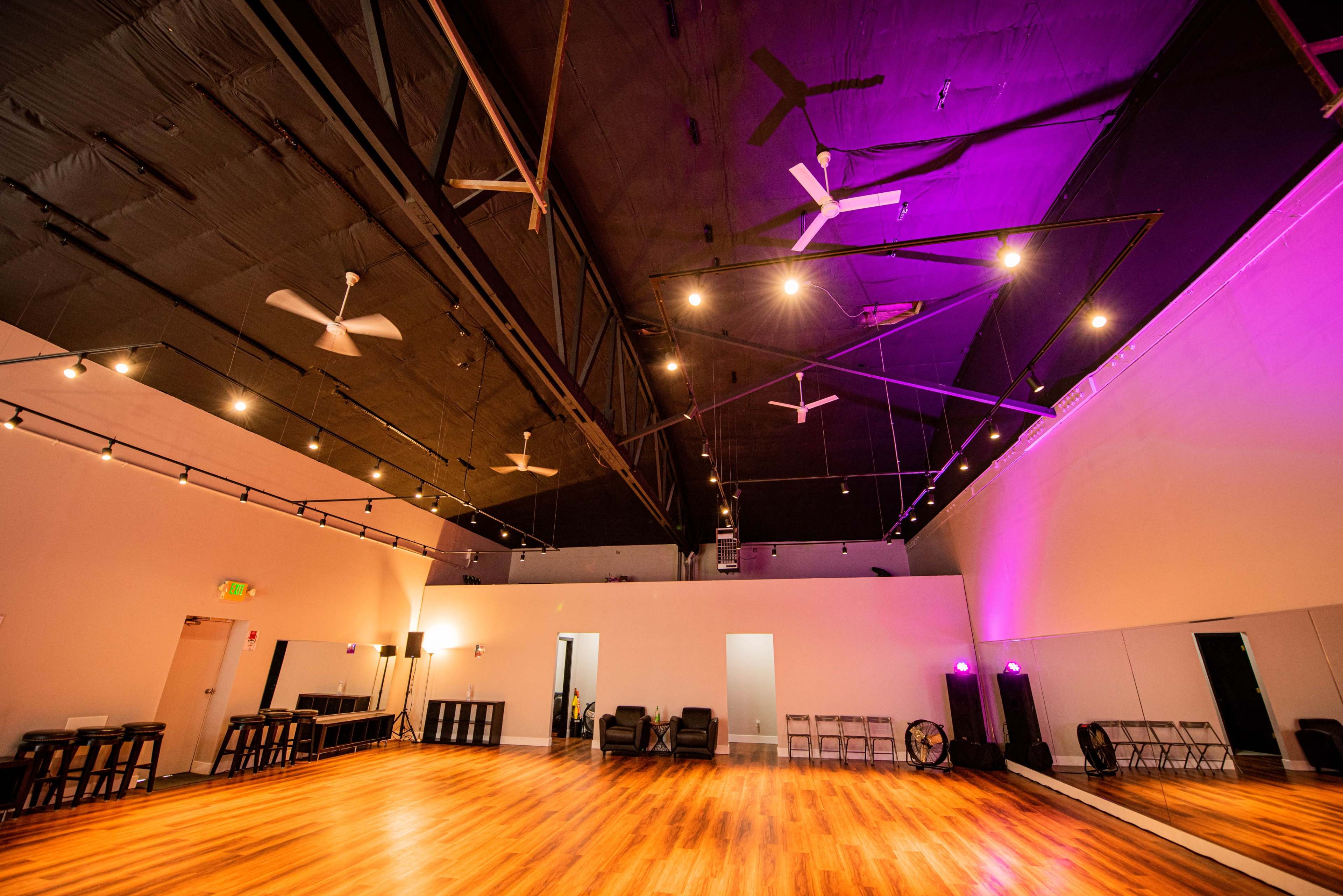 A spacious dance studio with wooden flooring, a high ceiling, and multiple ceiling fans, illuminated by purple and white lights.