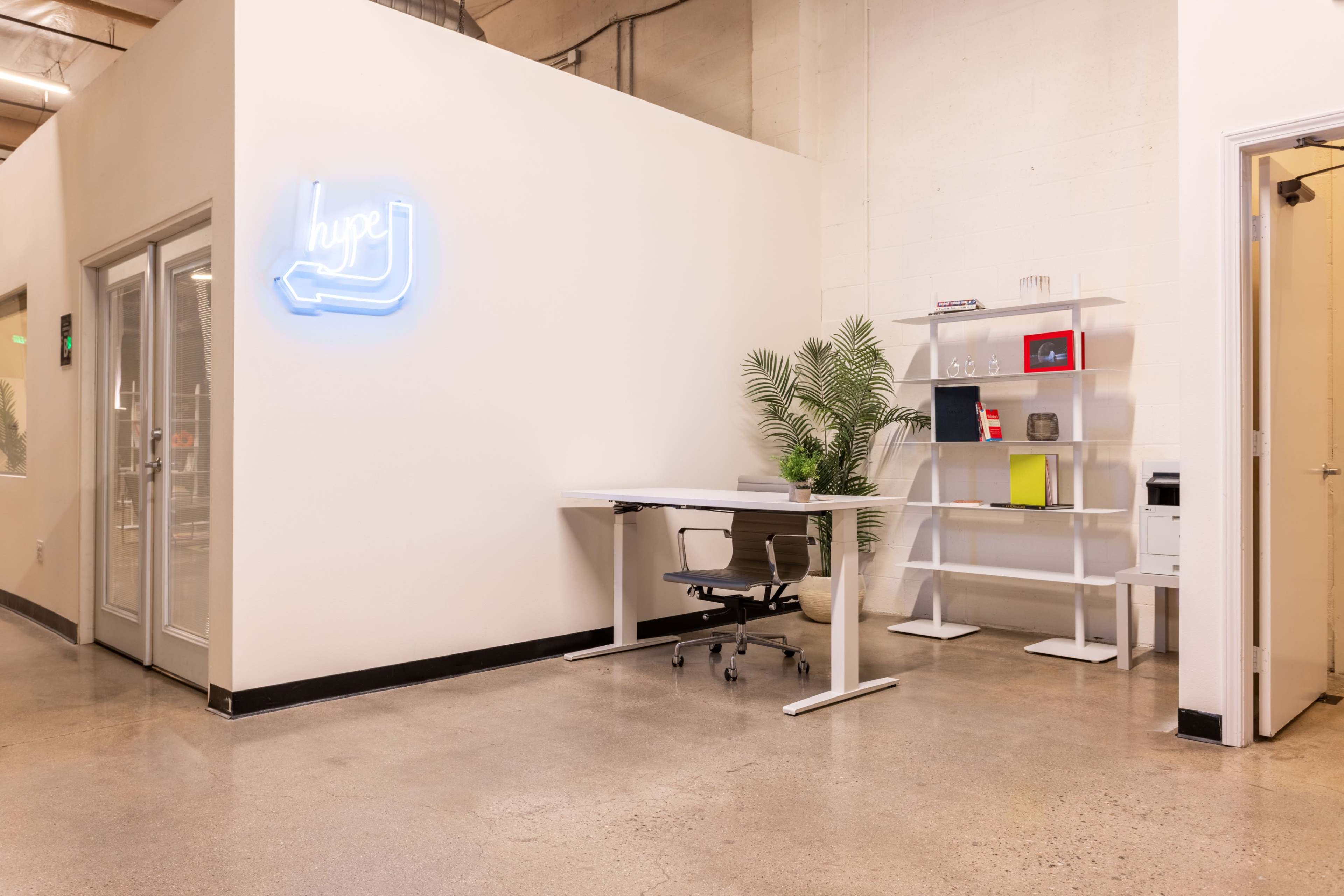 A modern office space featuring a desk with an office chair, a shelving unit filled with various items, and a neon sign that says "hype."