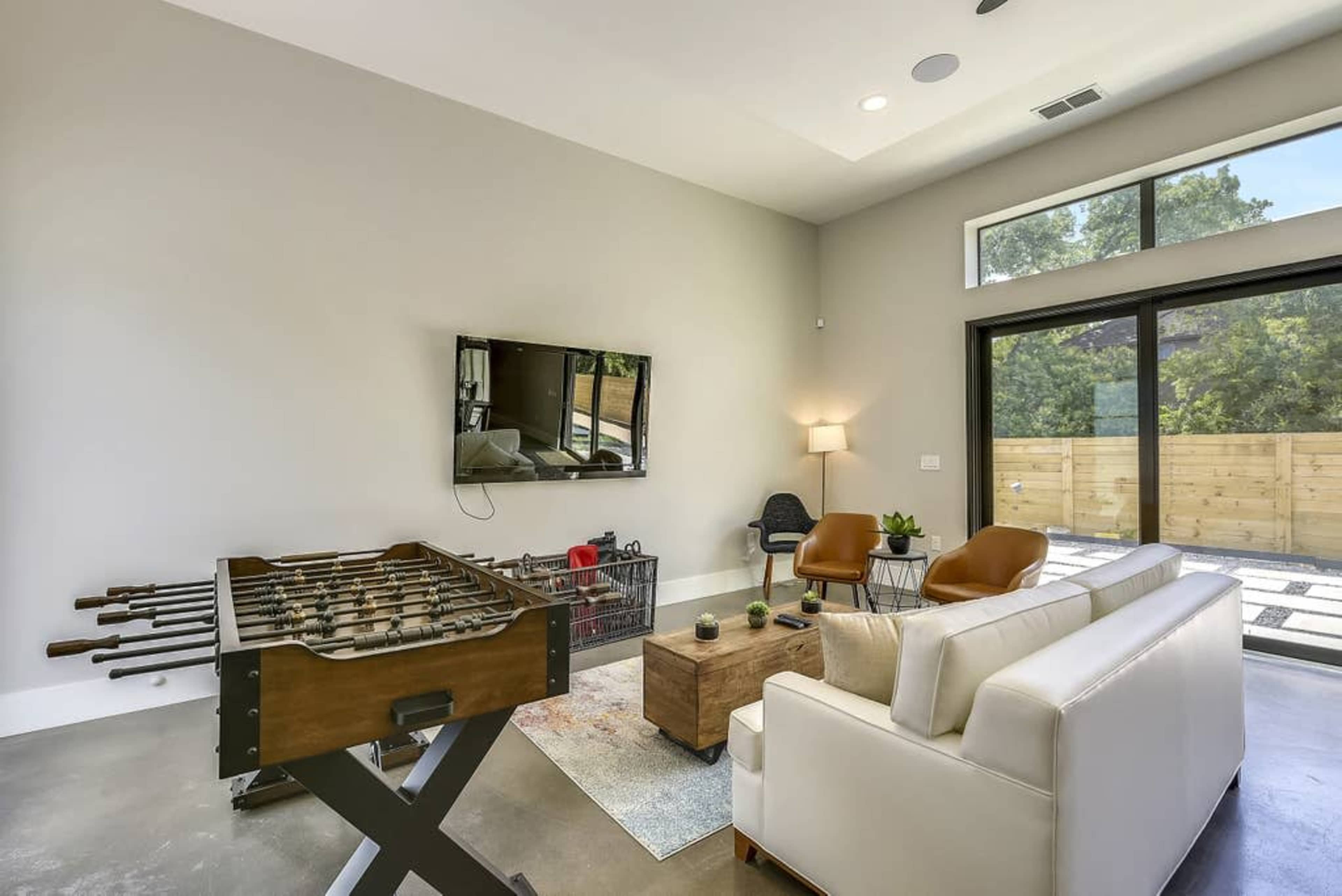 A contemporary living room featuring a foosball table, a white sofa, a coffee table, and a wall-mounted television, with large windows providing natural light.