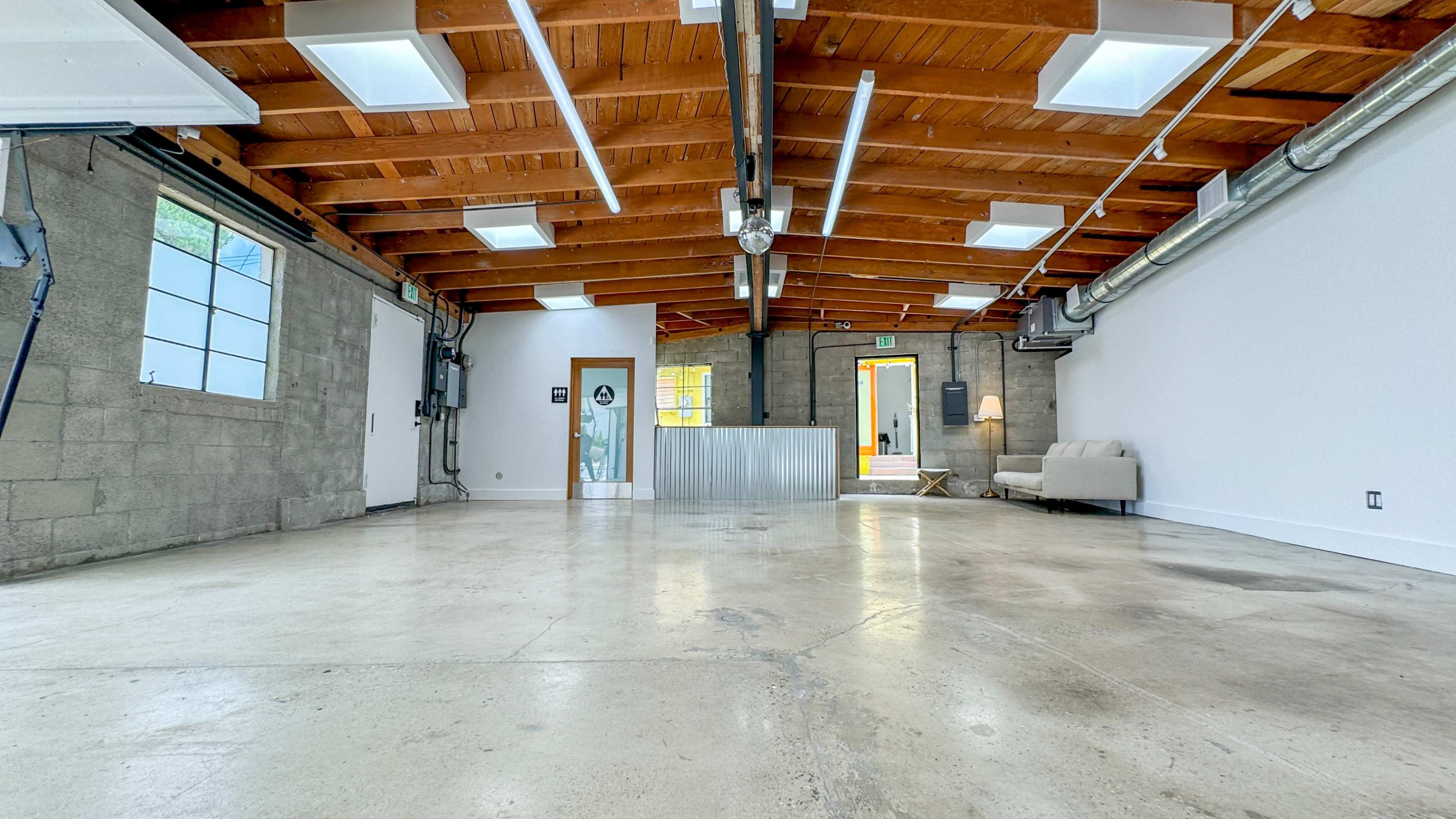 An expansive, empty room with a concrete floor, wooden ceiling beams, and large windows, featuring a desk and a couch against the walls.