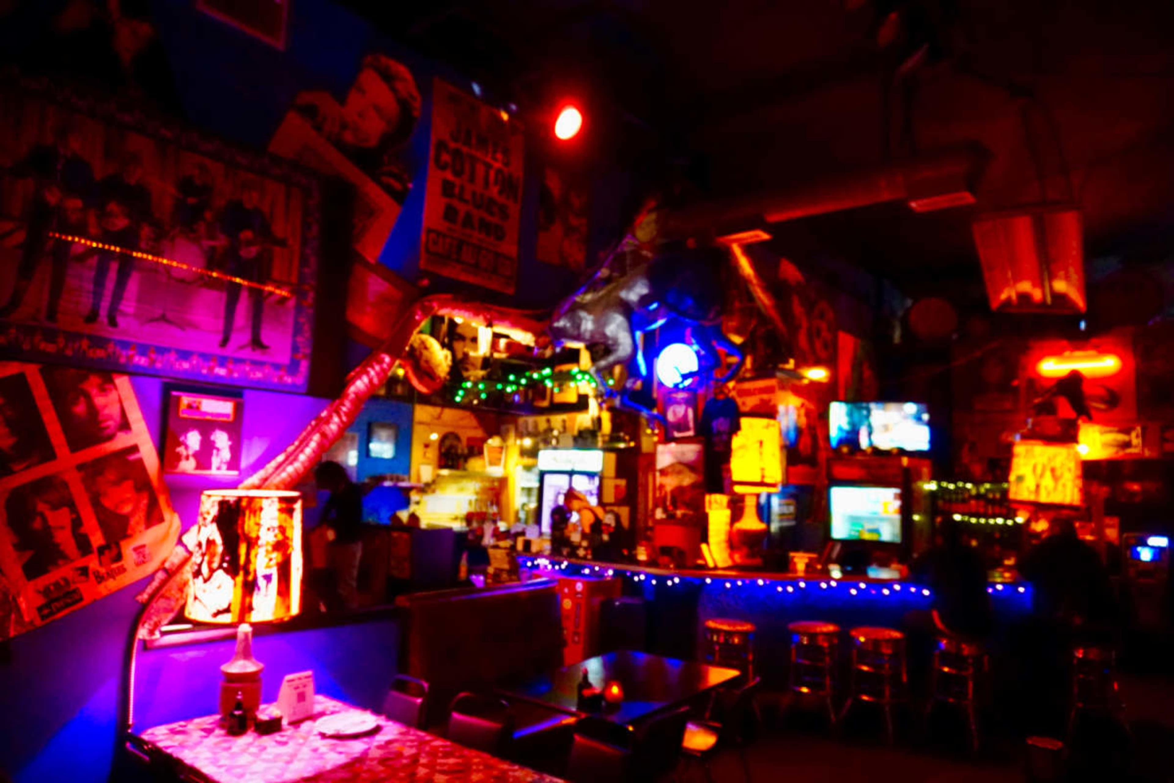 The image shows a dimly lit bar decorated with colorful neon lights, vintage posters, and a lively atmosphere.