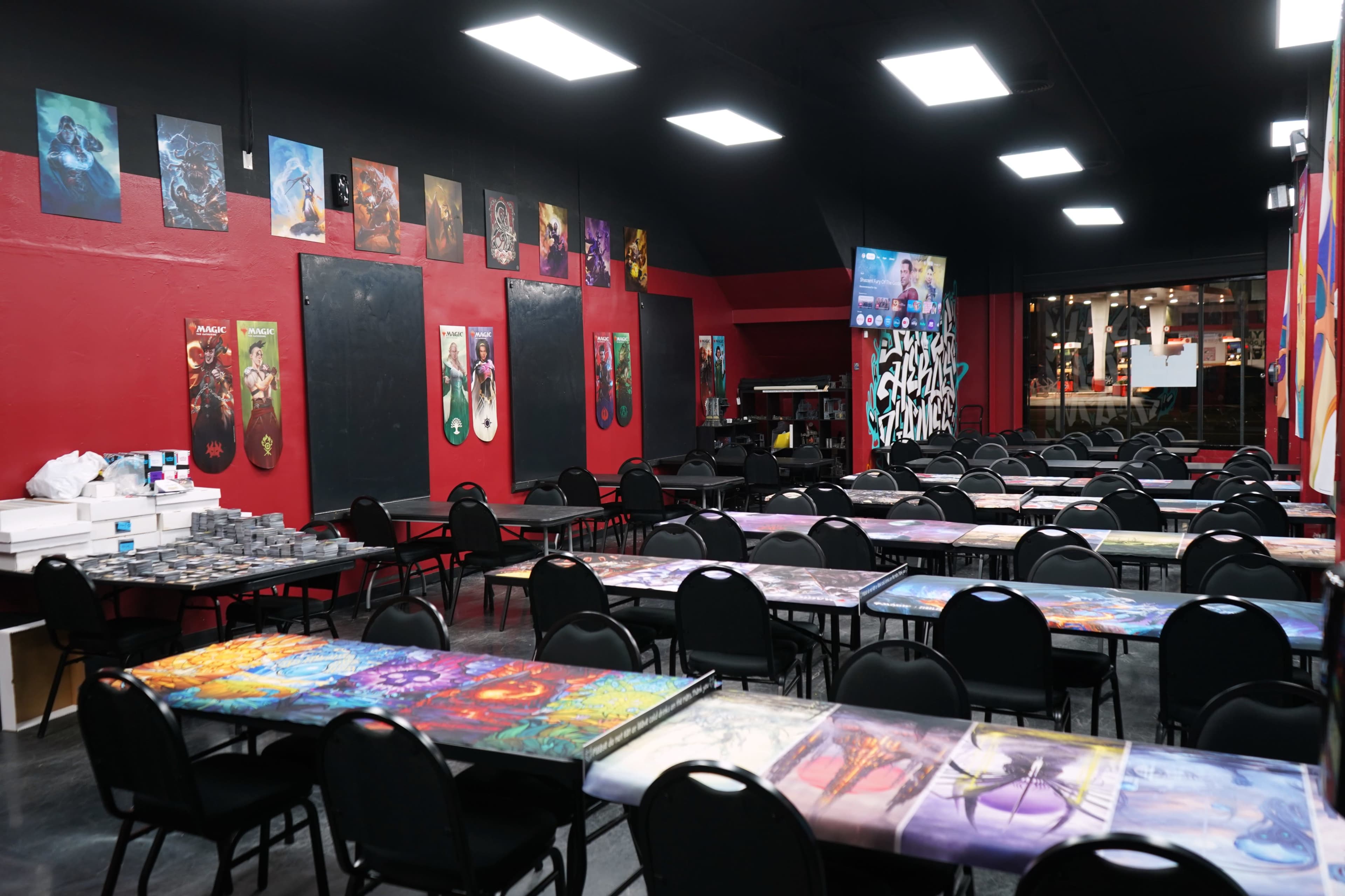 The image shows a gaming venue with rows of tables covered in colorful art and walls adorned with posters.