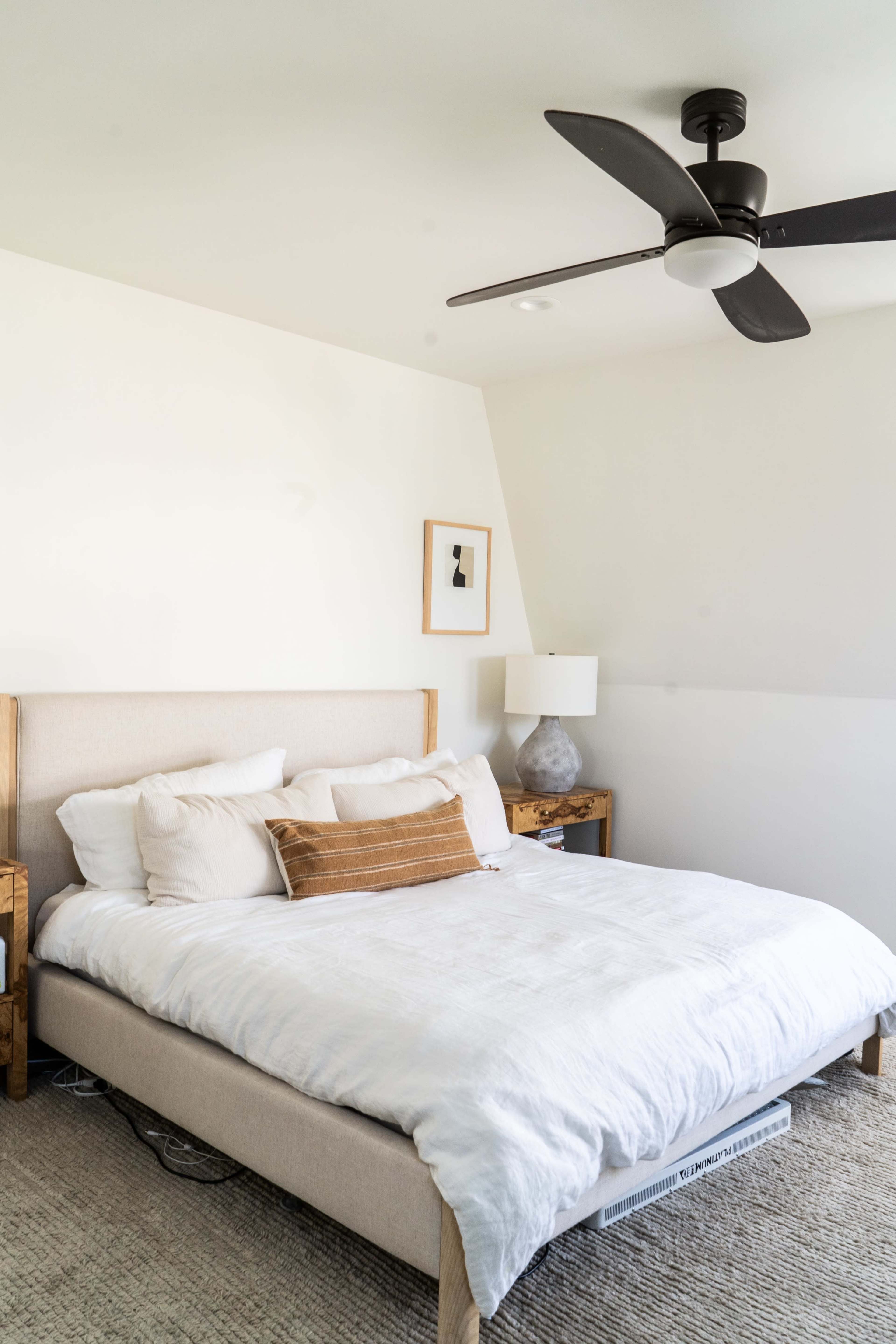 The image shows a neatly made bed with a light-colored comforter in a minimalist bedroom featuring a fan and a piece of wall art.