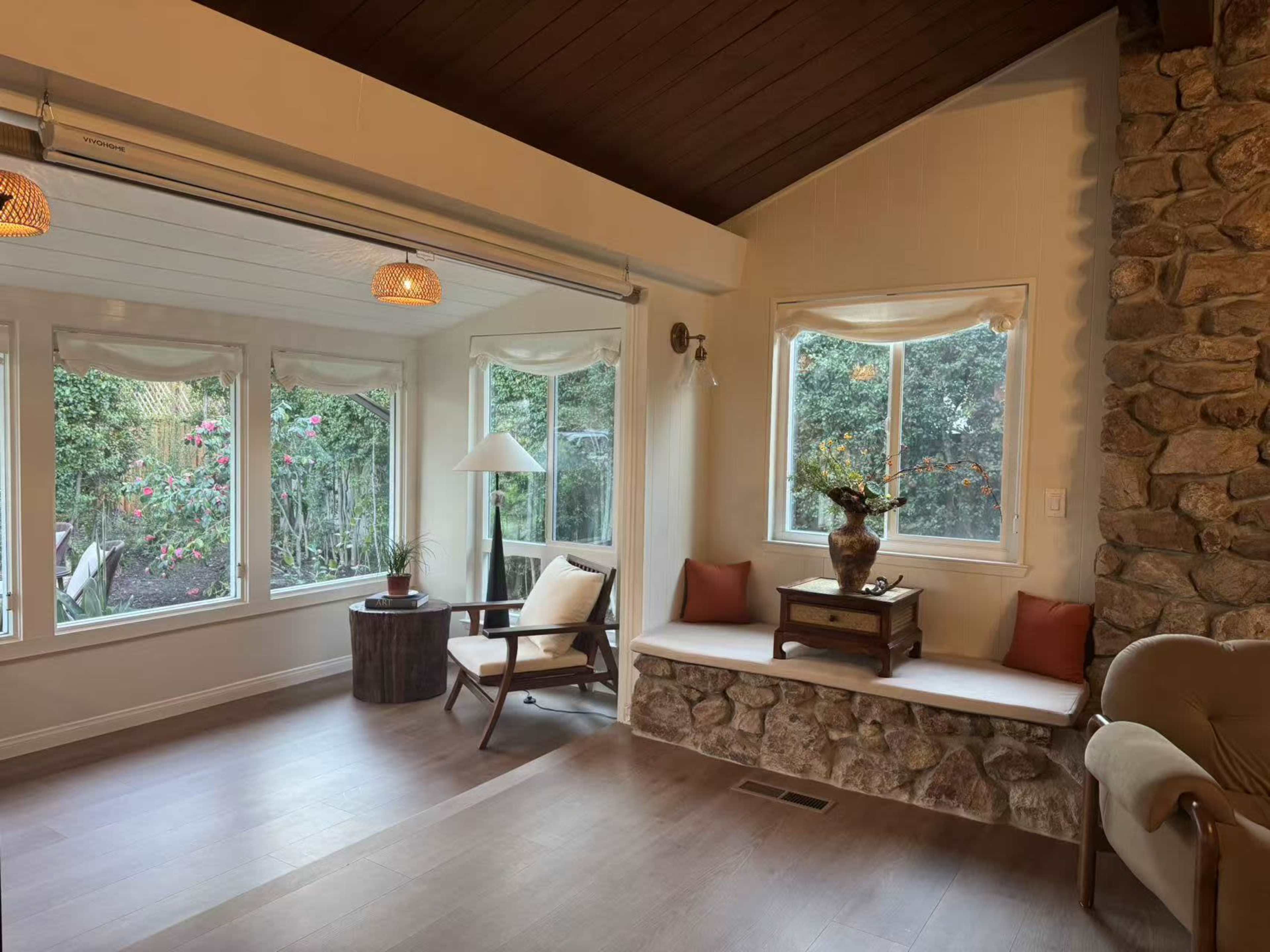 The image shows a cozy sunroom with large windows, a stone bench, and comfortable seating arrangements.