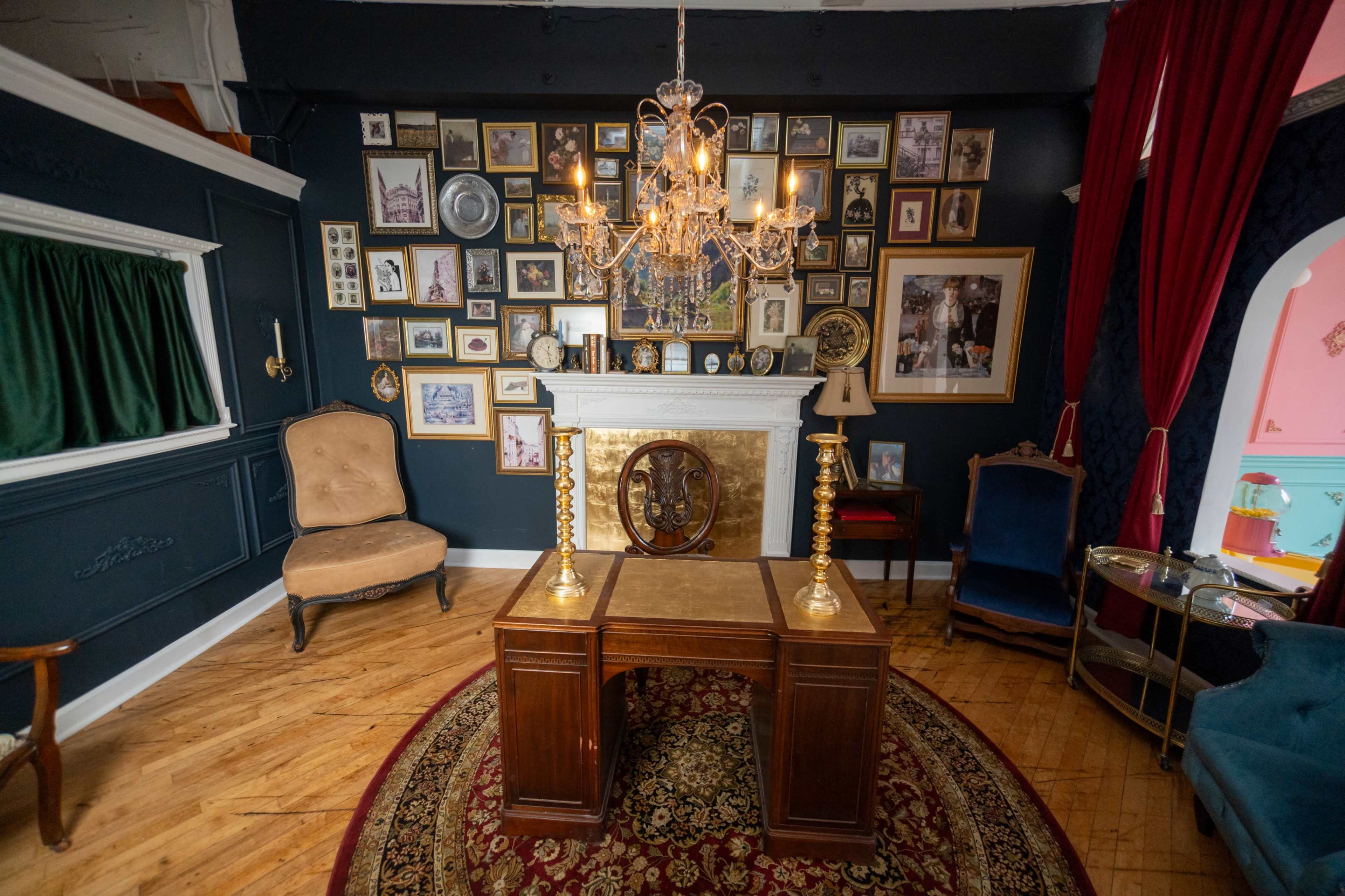 The image shows a room with dark walls, a decorative chandelier hanging from the ceiling, a wooden table in the center, and a variety of framed pictures arranged on the wall behind a fireplace.