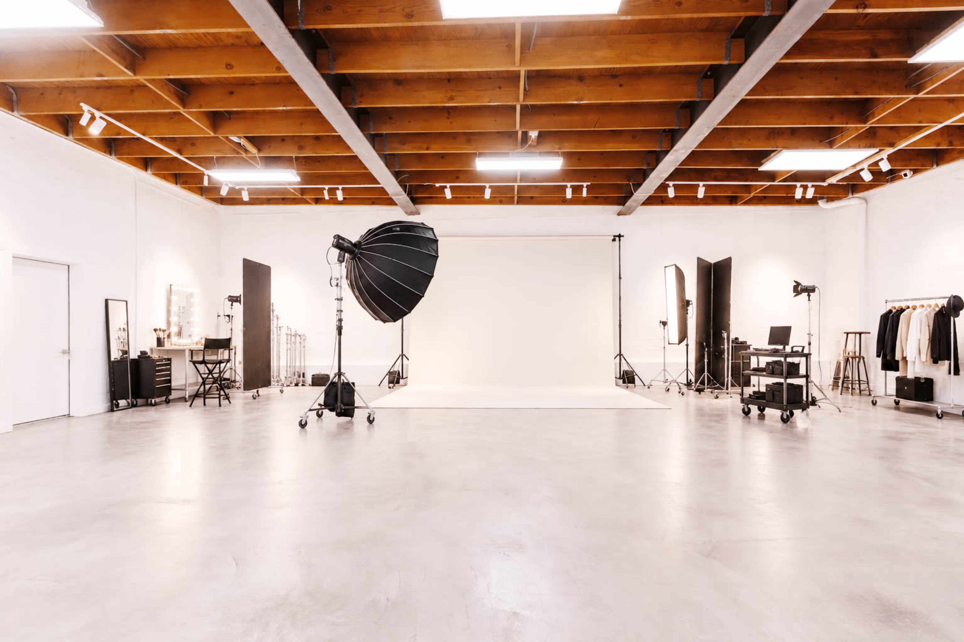 The image shows a spacious photography studio with a plain backdrop, lighting equipment, and various clothing items displayed on racks.