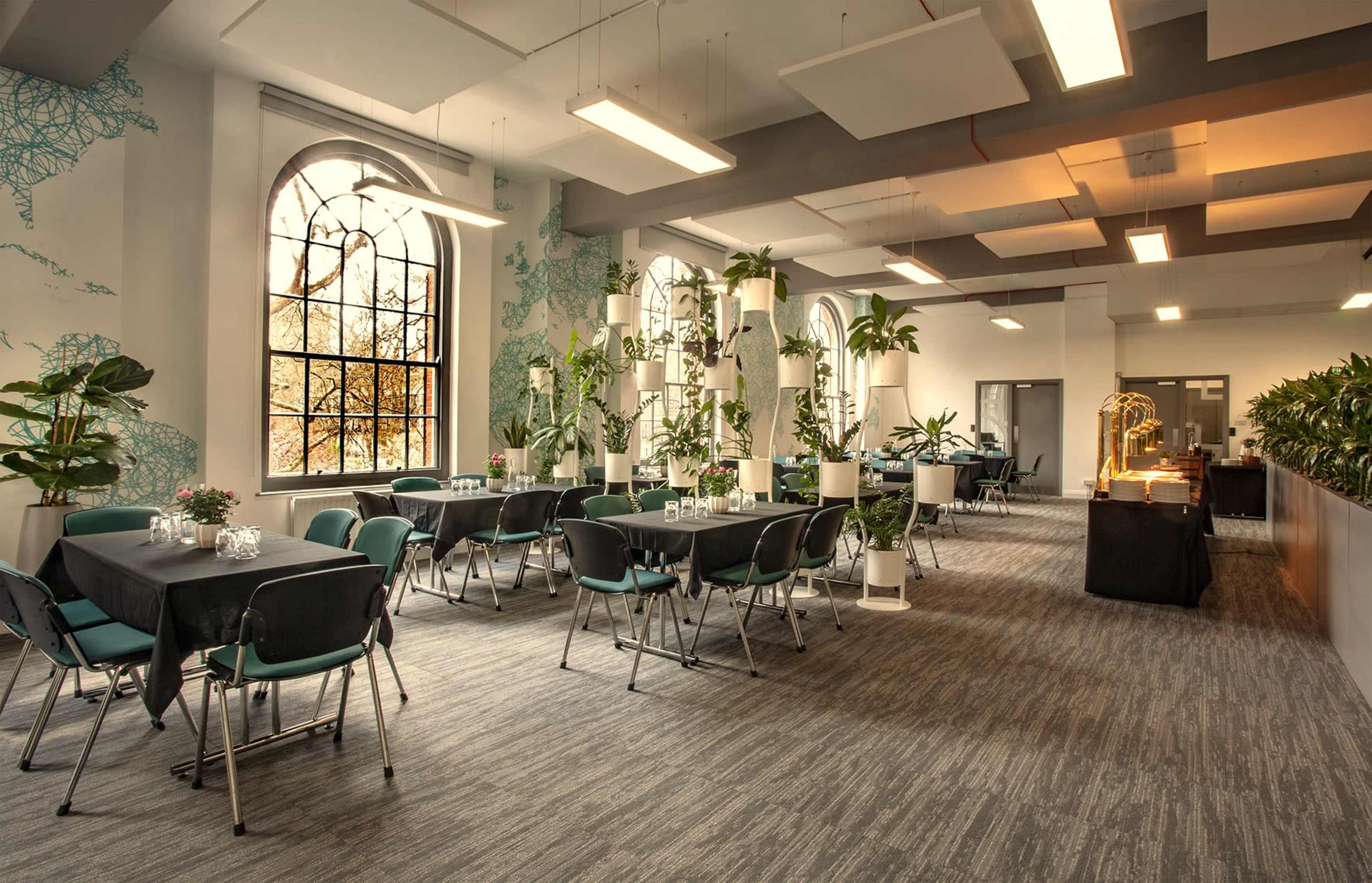 A large dining area with tables and chairs arranged among various potted plants and large windows.