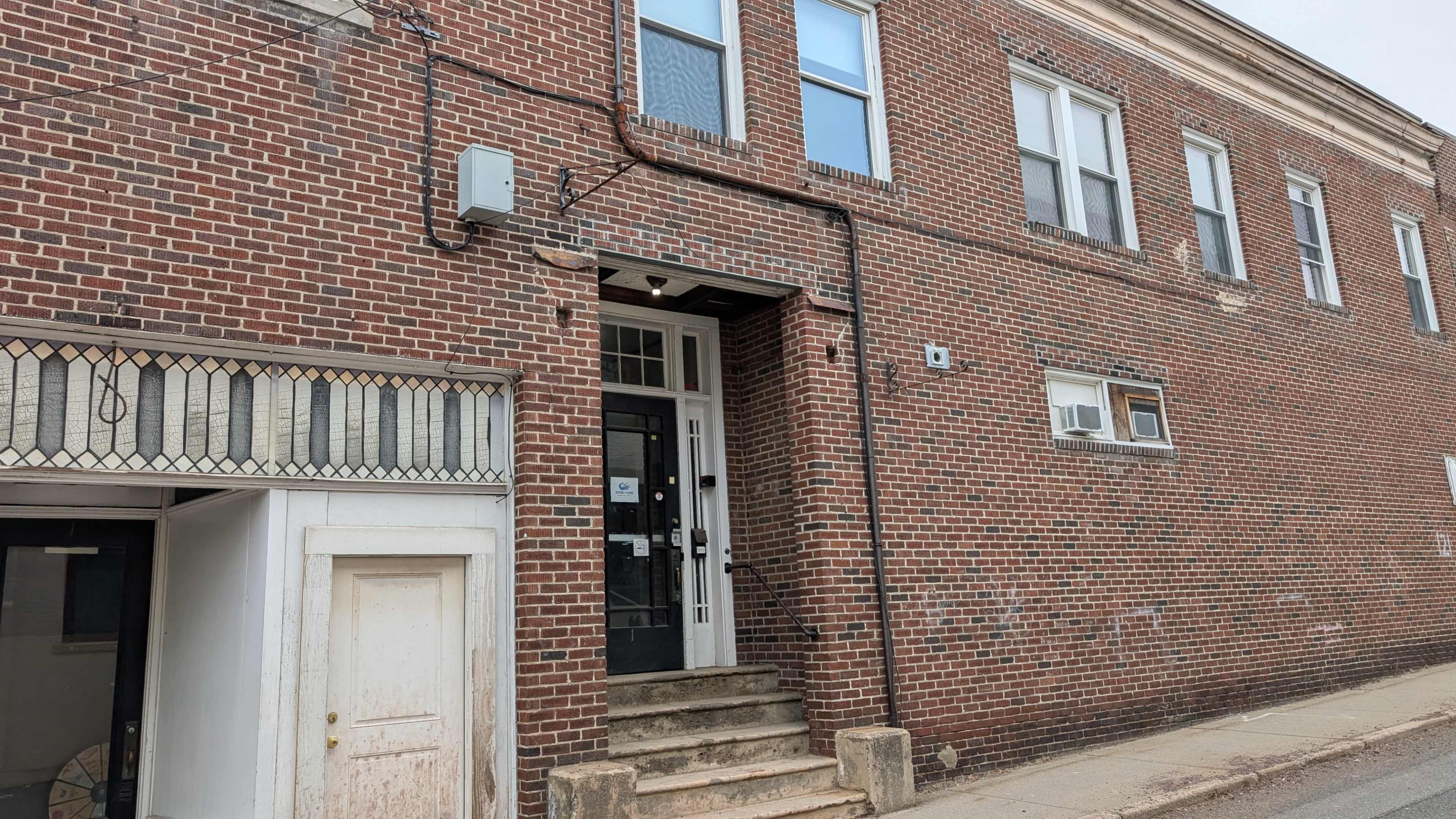 The image shows a brick building with a staircase leading to a door, alongside a smaller white structure on the left.