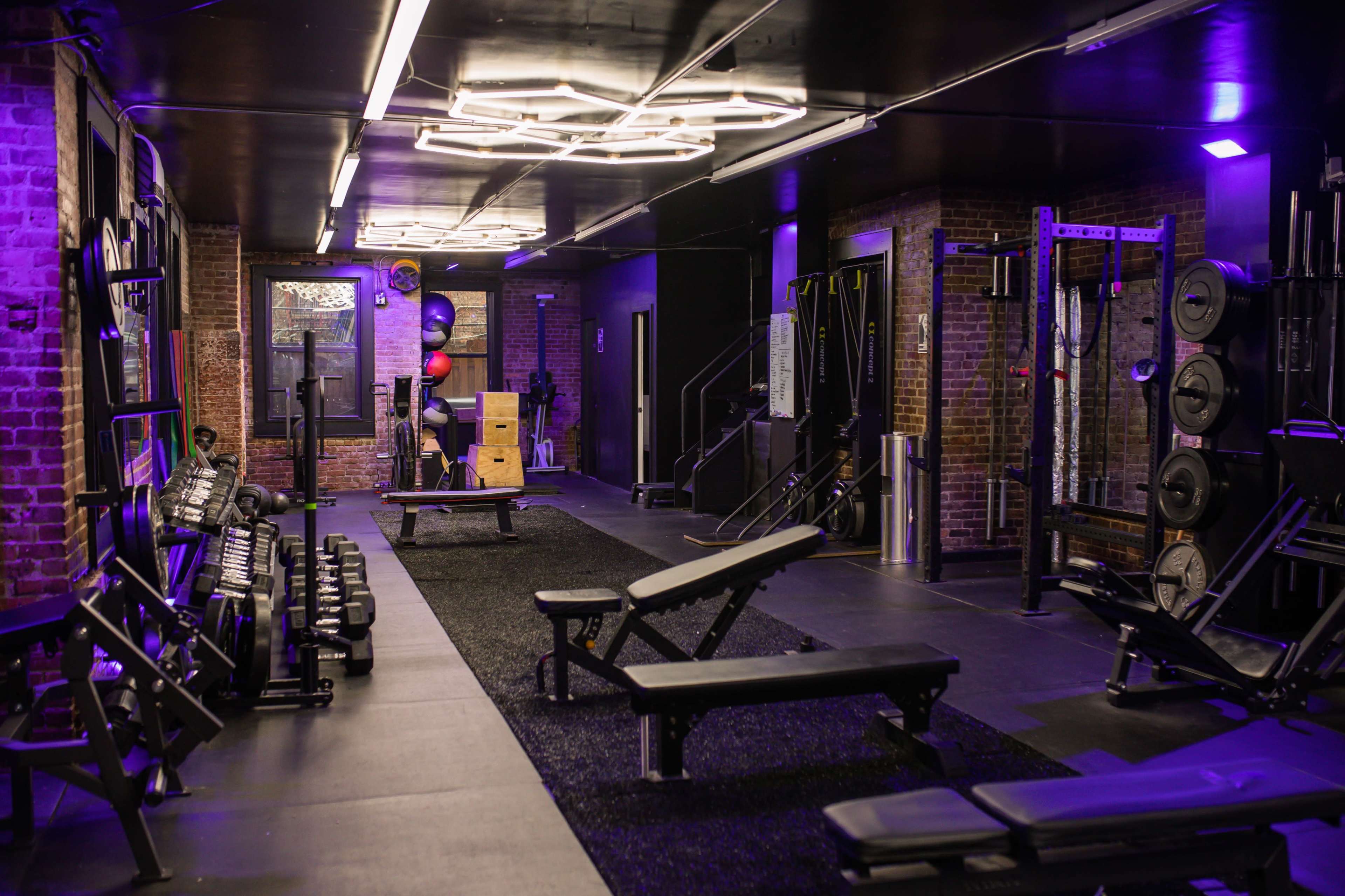 The image shows a well-equipped gym featuring dumbbells, benches, and various workout stations under purple lighting.