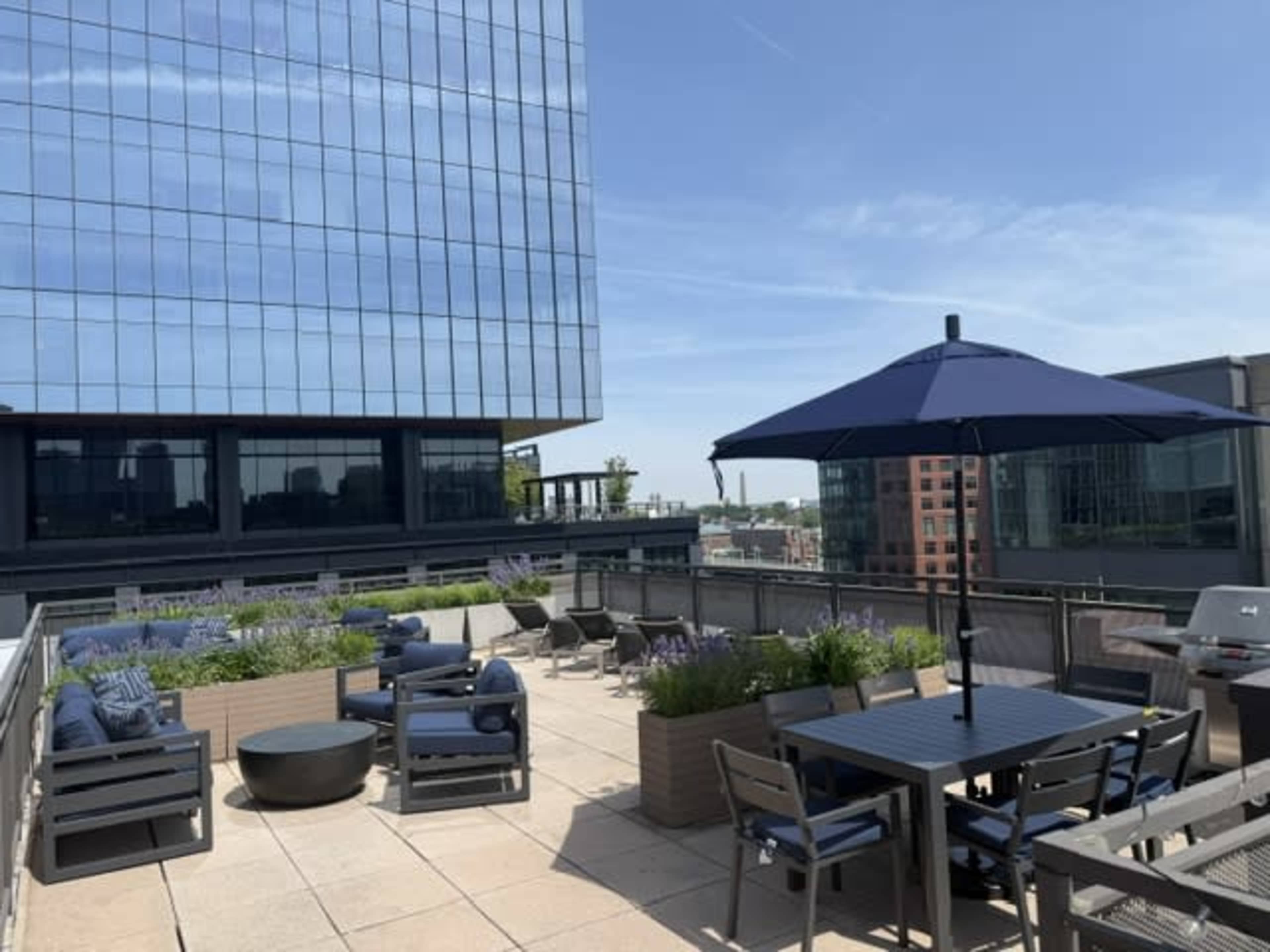 The image depicts a rooftop terrace with seating areas, a table, and an umbrella, surrounded by modern buildings under a clear blue sky.