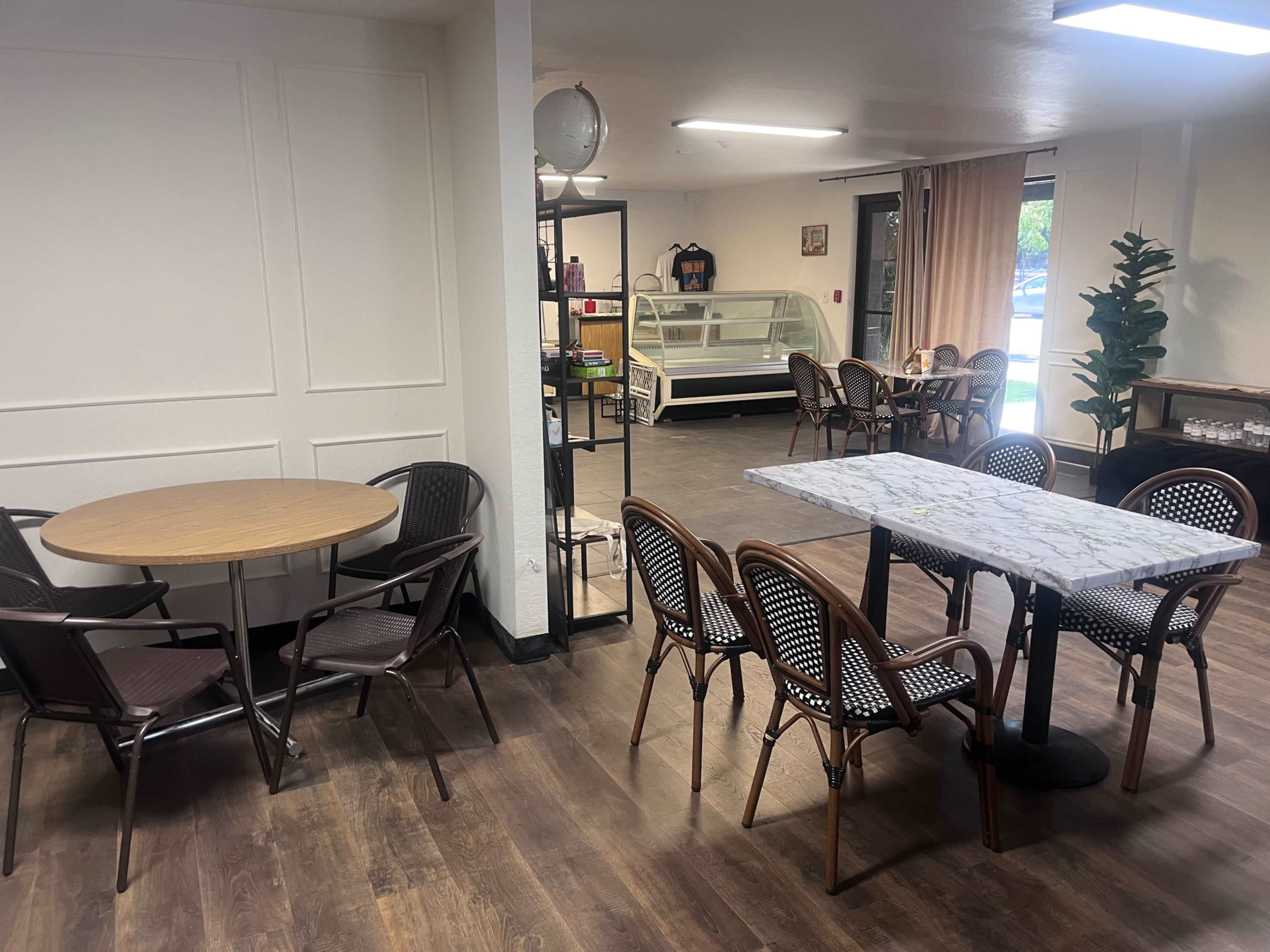 The image shows a spacious indoor setting with wooden flooring, featuring two dining areas: one with round tables and wicker chairs, and another with a rectangular table and additional seating.