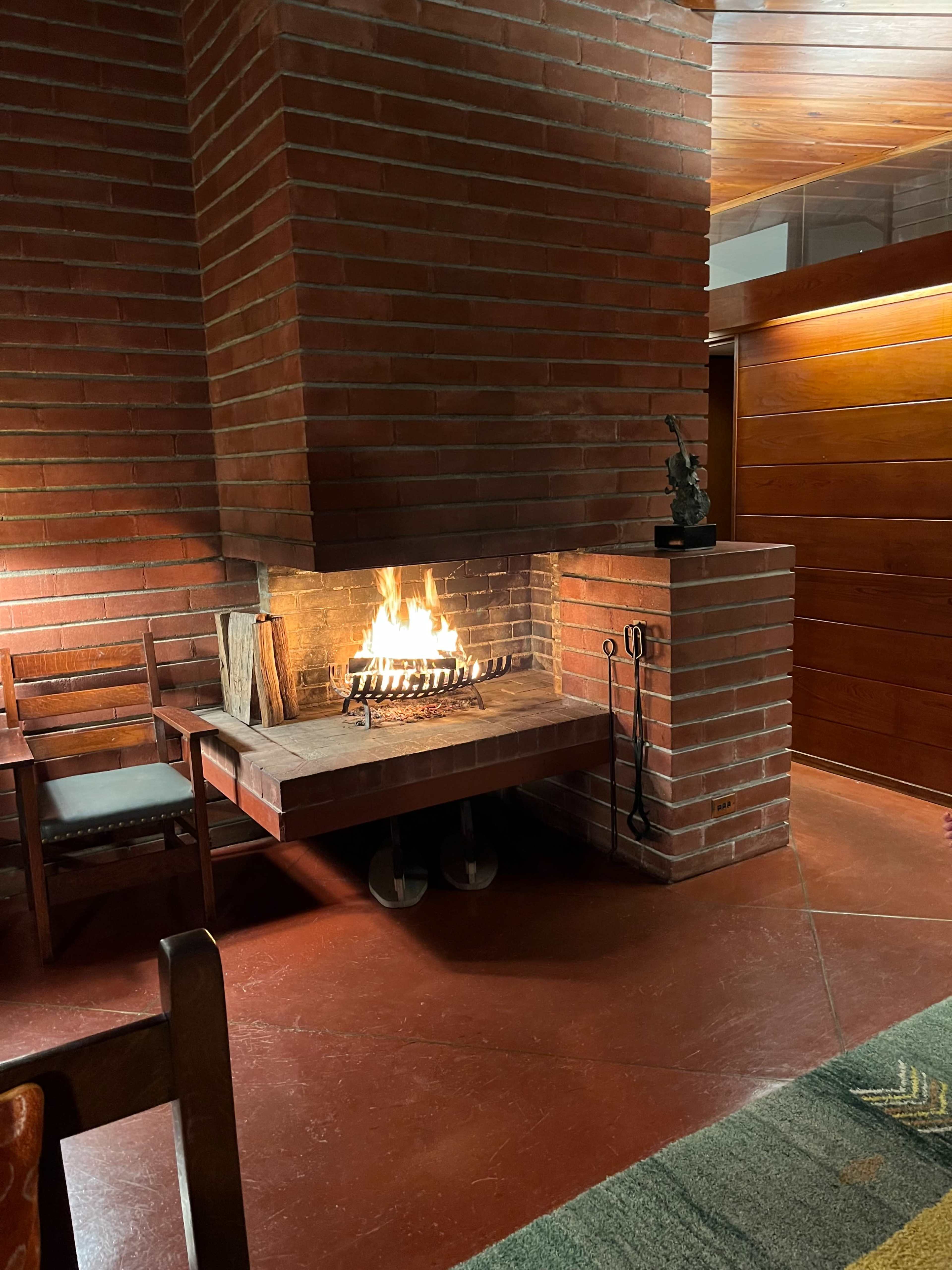 A brick fireplace with a lit fire is positioned in a cozy room featuring wooden chairs and a sculpture.
