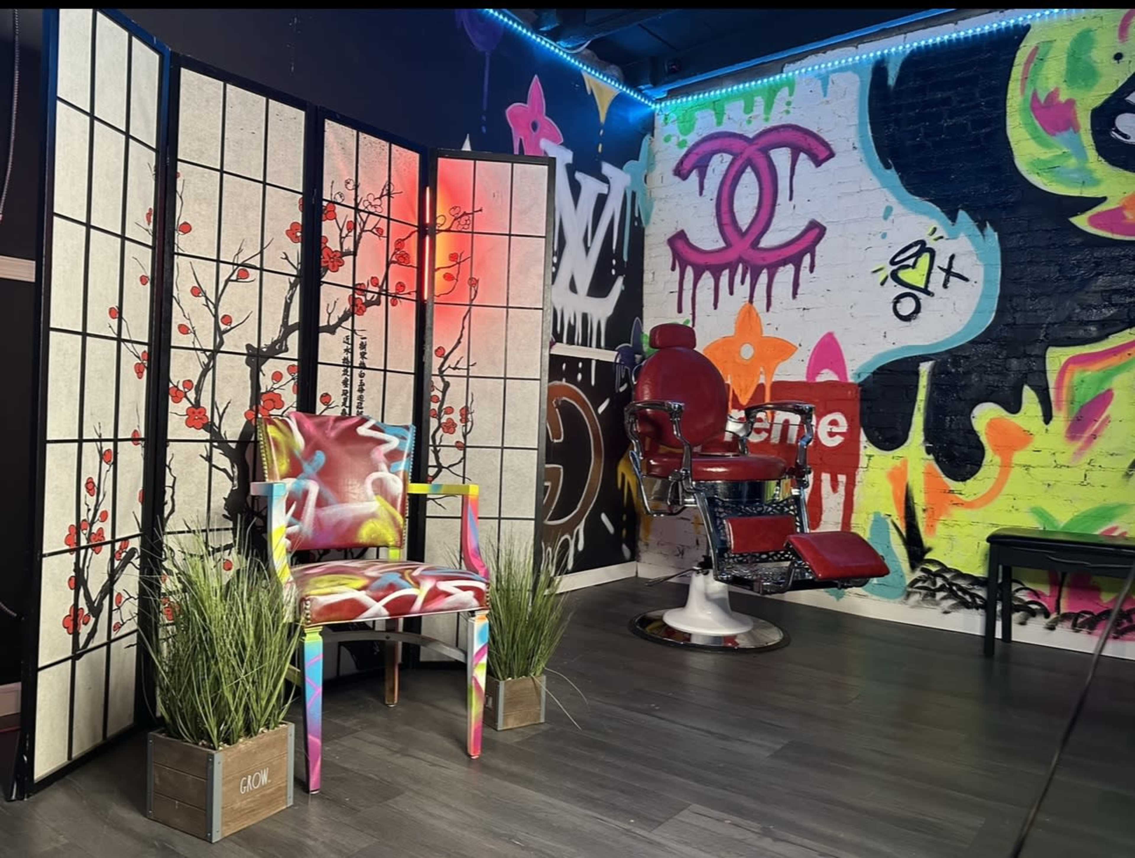 The image shows a vibrant tattoo studio featuring a colorful chair, a folding screen with floral motifs, and a barber's chair against a graffiti-covered wall.