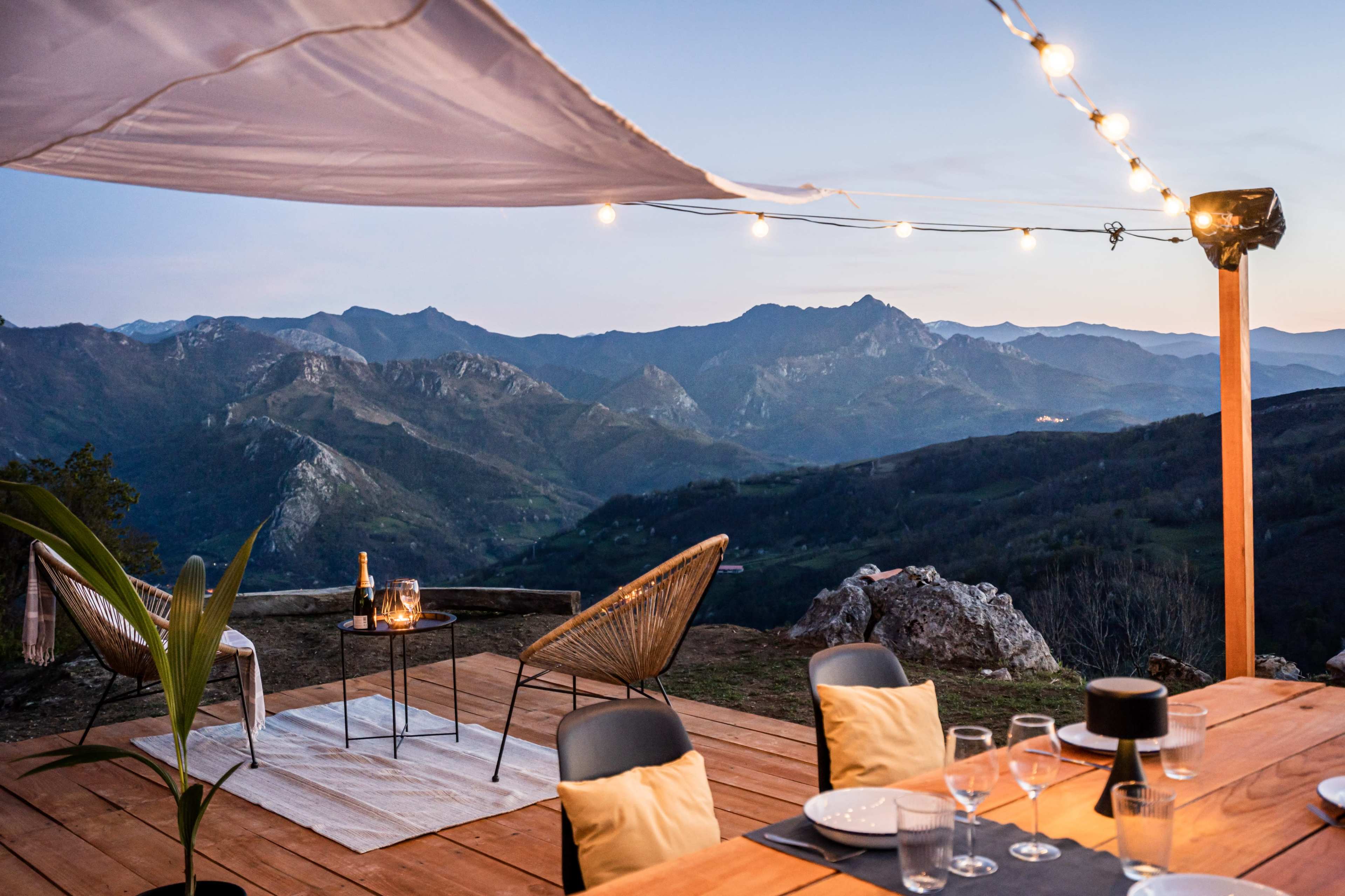 A wooden deck features a table set for dinner, surrounded by chairs, with mountains visible in the background under a twinkling string of lights.