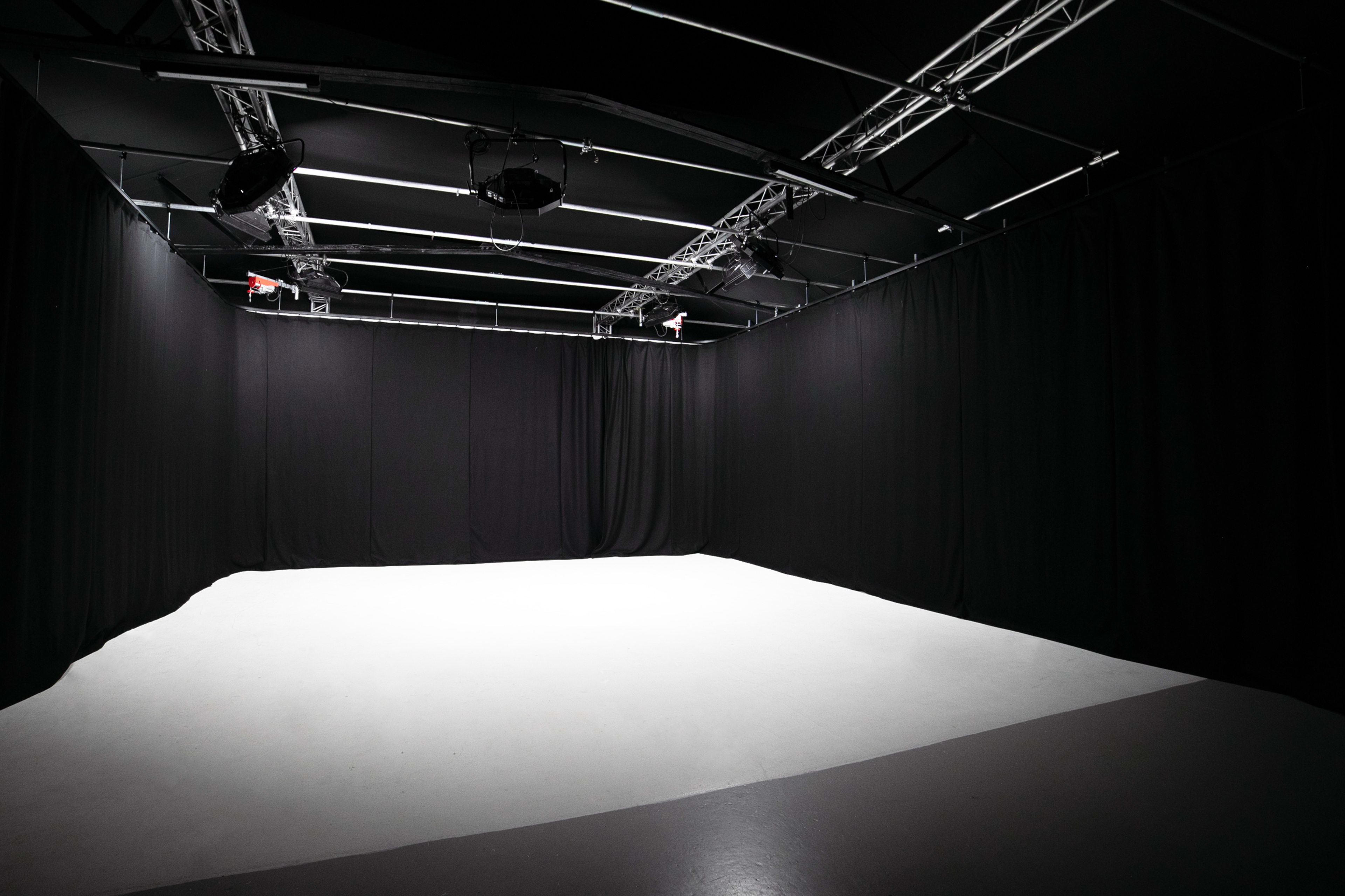 The image shows an empty studio space enclosed by black curtains and illuminated by overhead lights.