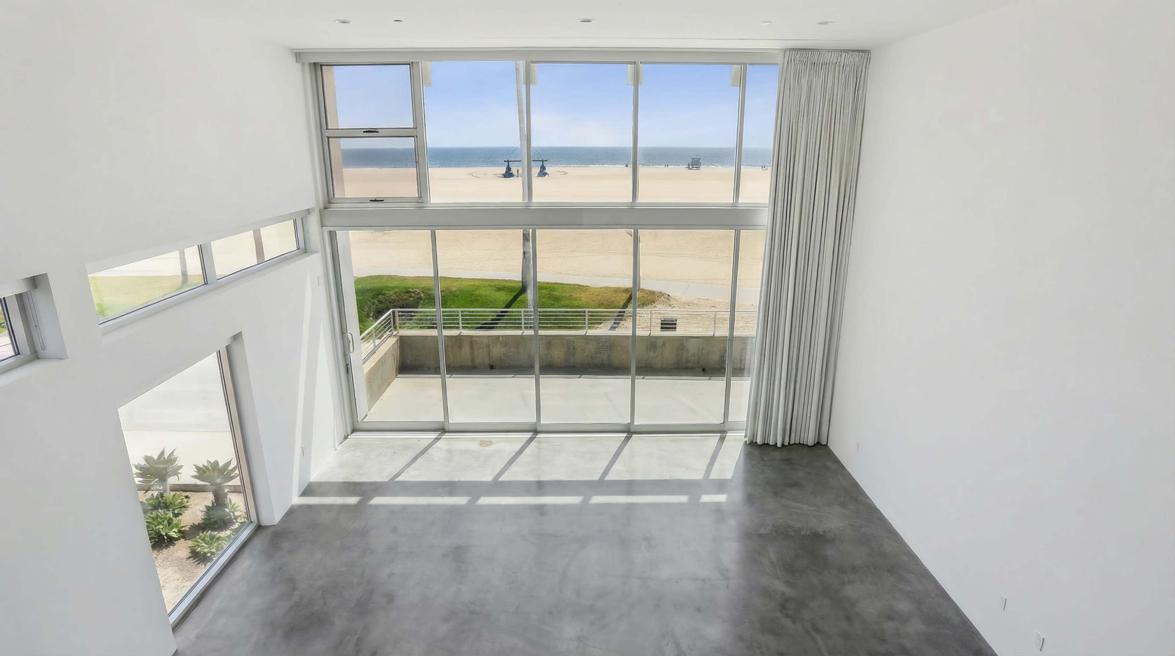 The image shows a spacious, modern interior with large glass windows overlooking a beach.