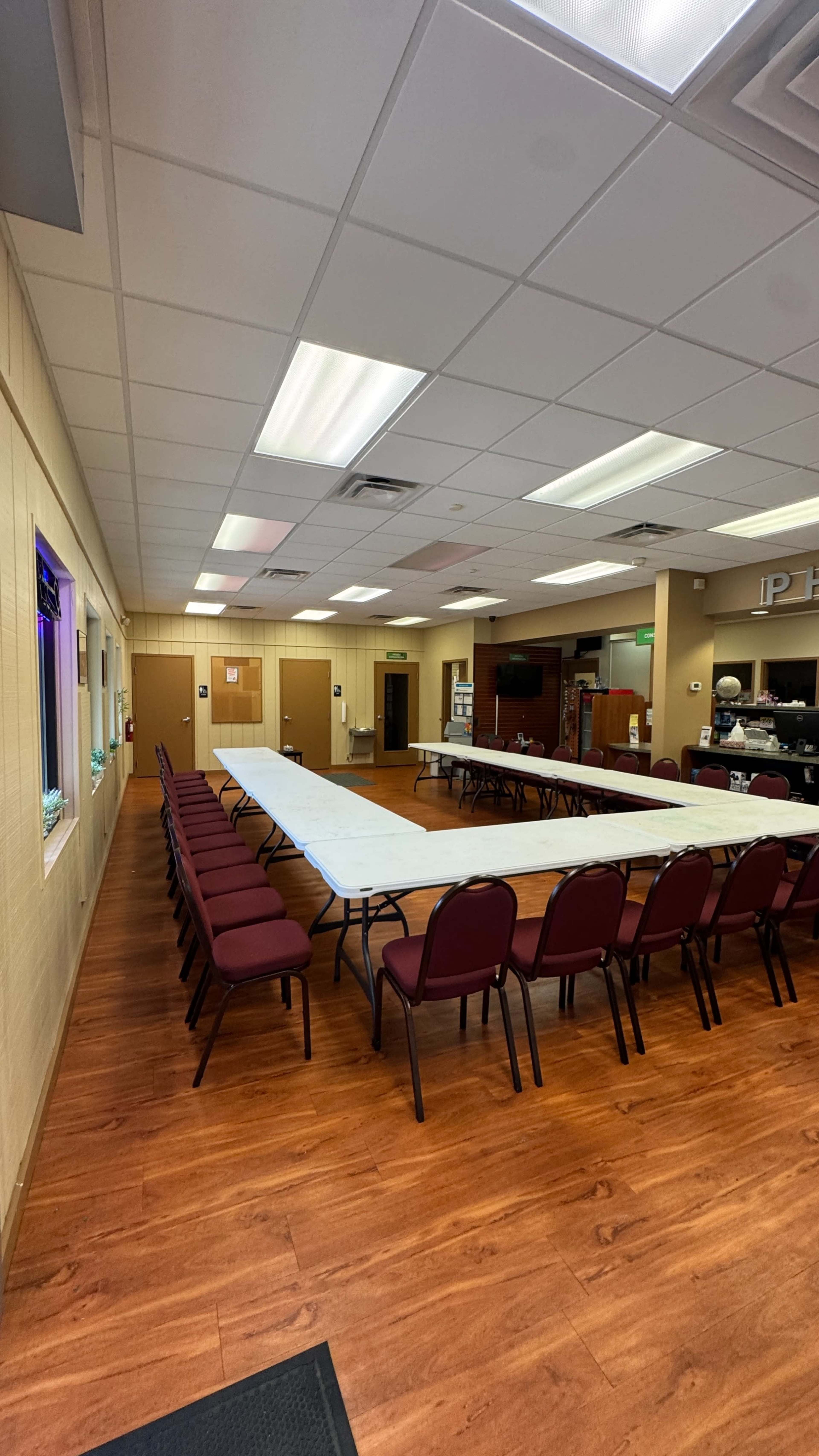 The image shows a spacious meeting room with long tables arranged in a U-shape and several chairs lined up around them, featuring a wooden floor and soft lighting.