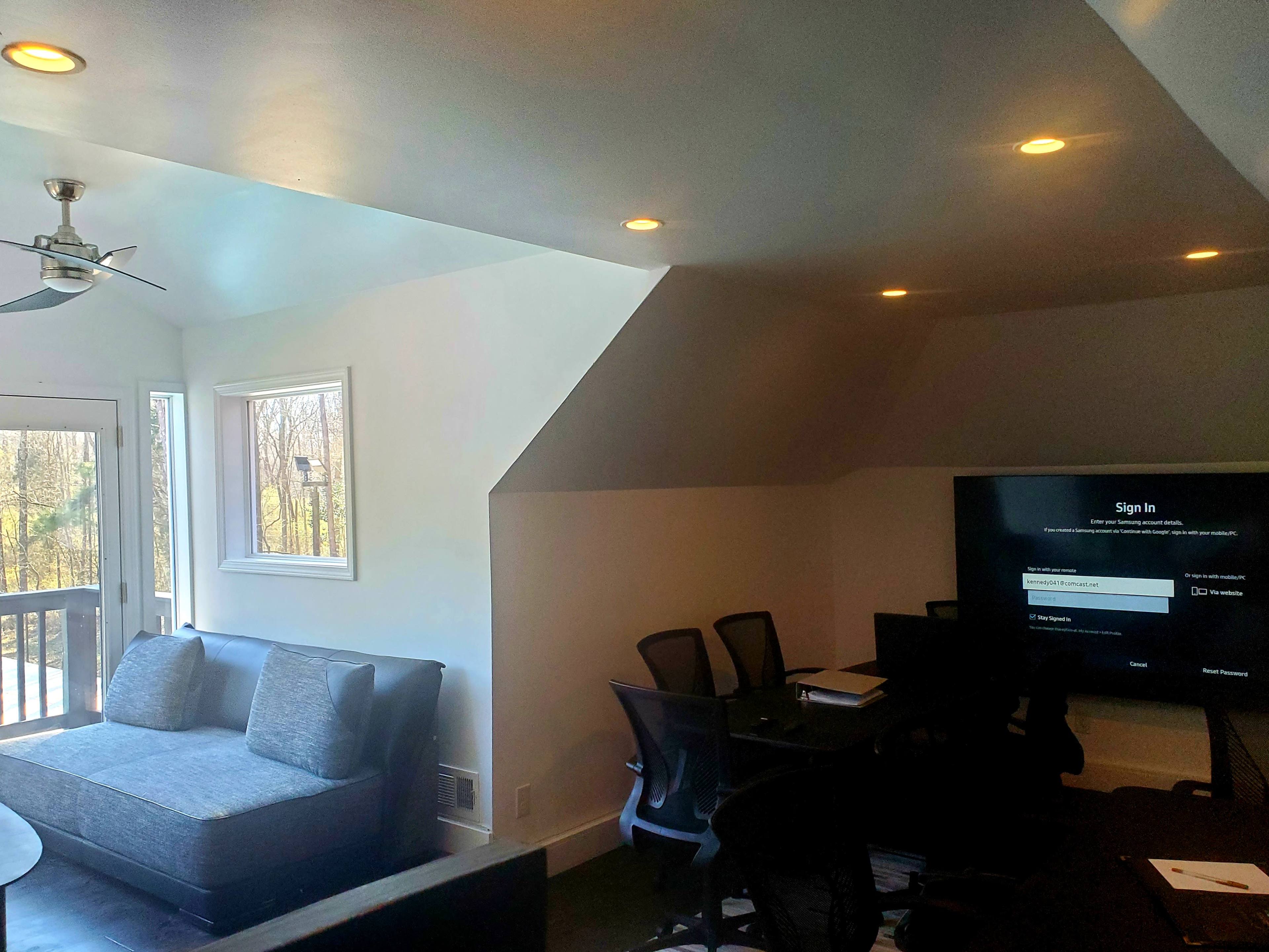 A modern living space with a gray sofa, a large television, and a table with chairs, illuminated by recessed lighting.