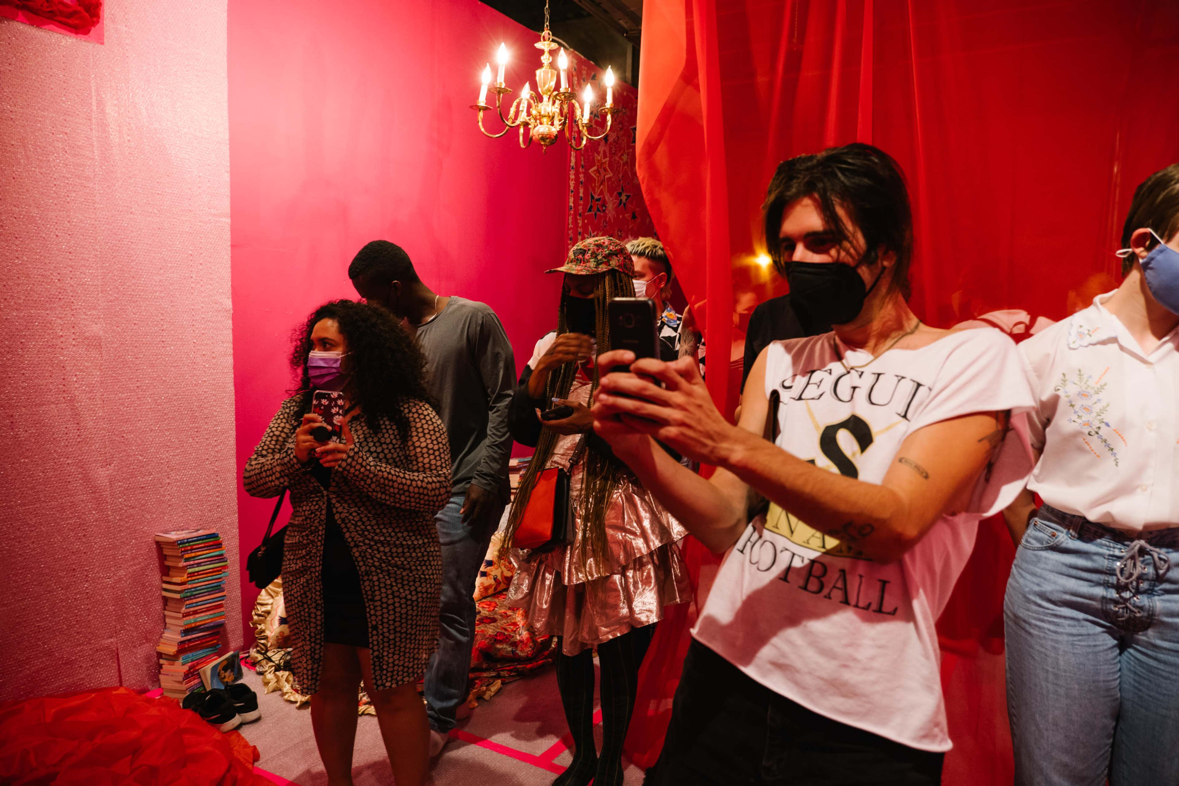 A group of people wearing masks are taking photos in a dimly lit, pink-themed room adorned with fabric and a chandelier.