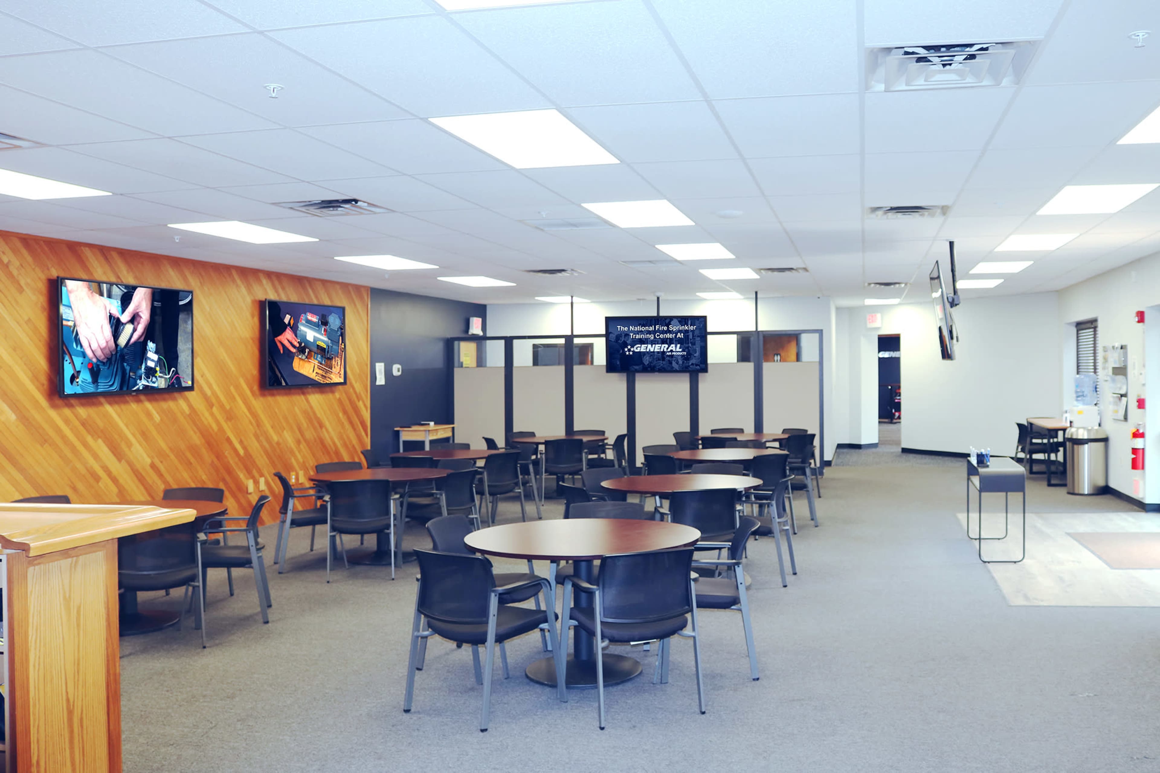 A spacious, well-lit room with several round tables and chairs, flanked by screens displaying various images on the walls.