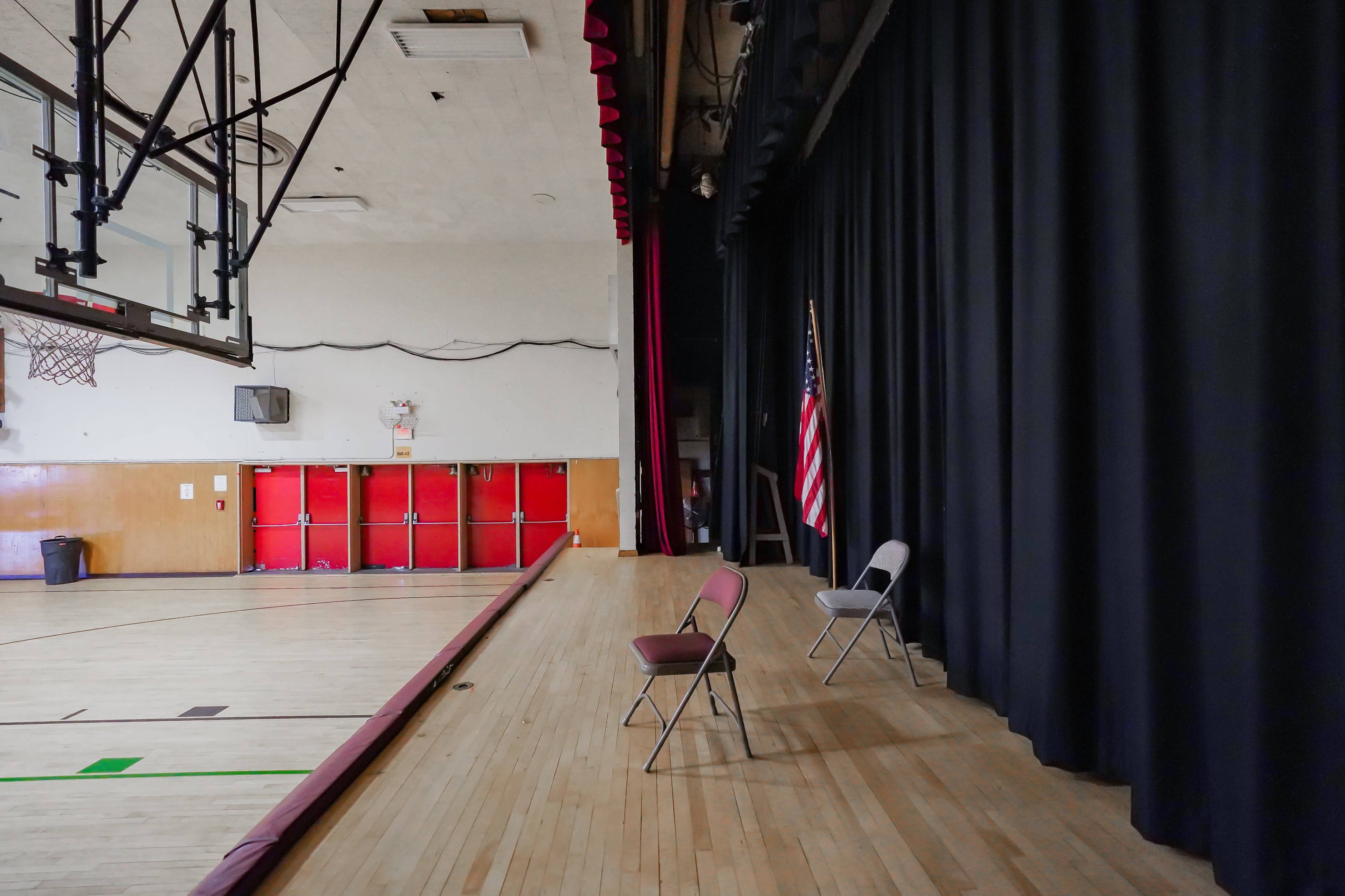The image shows a spacious gymnasium with two empty chairs positioned on a stage beside a black curtain and an American flag.