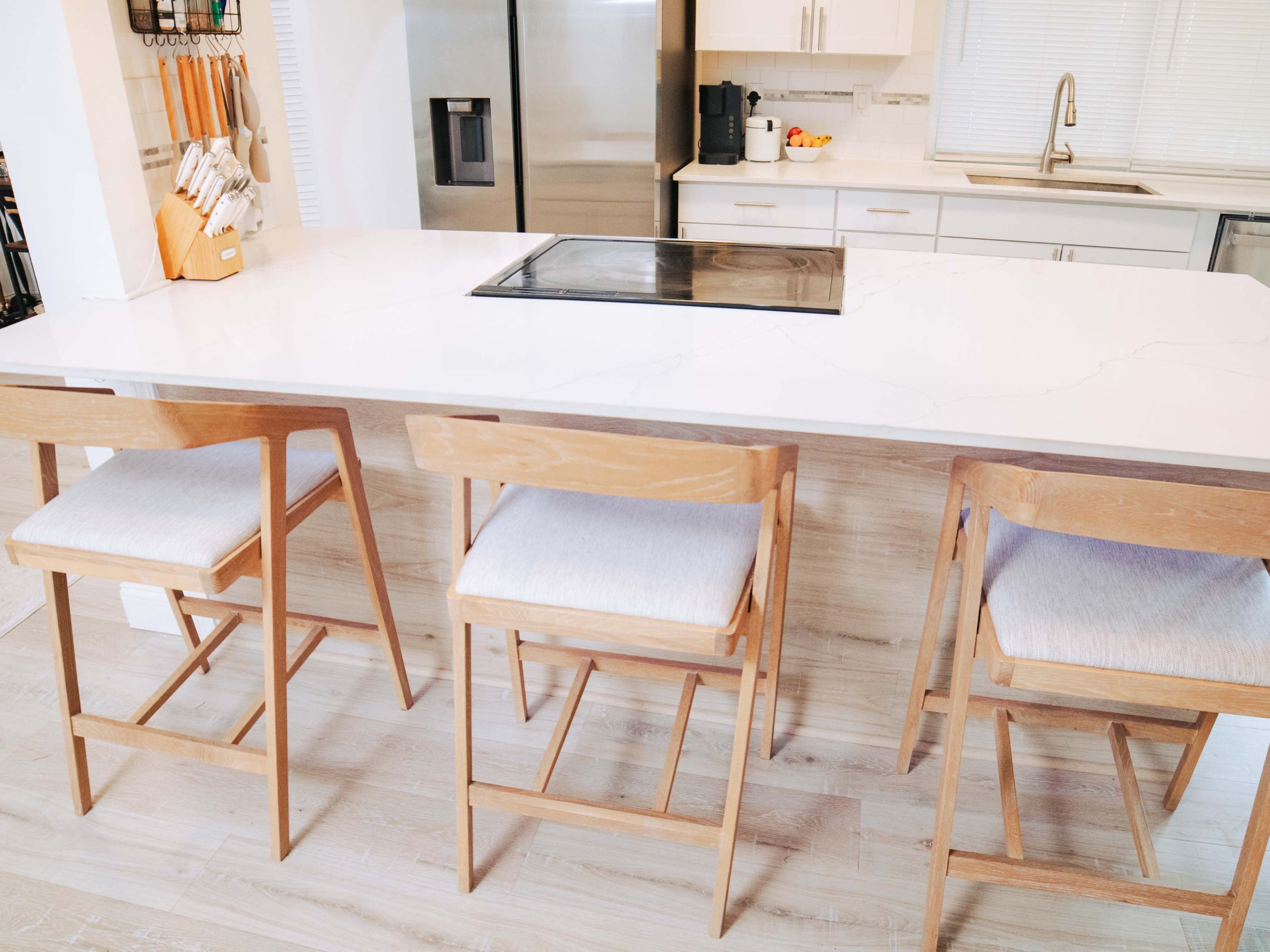 The image shows a modern kitchen island with three wooden bar stools and a stovetop embedded in a white marble countertop.