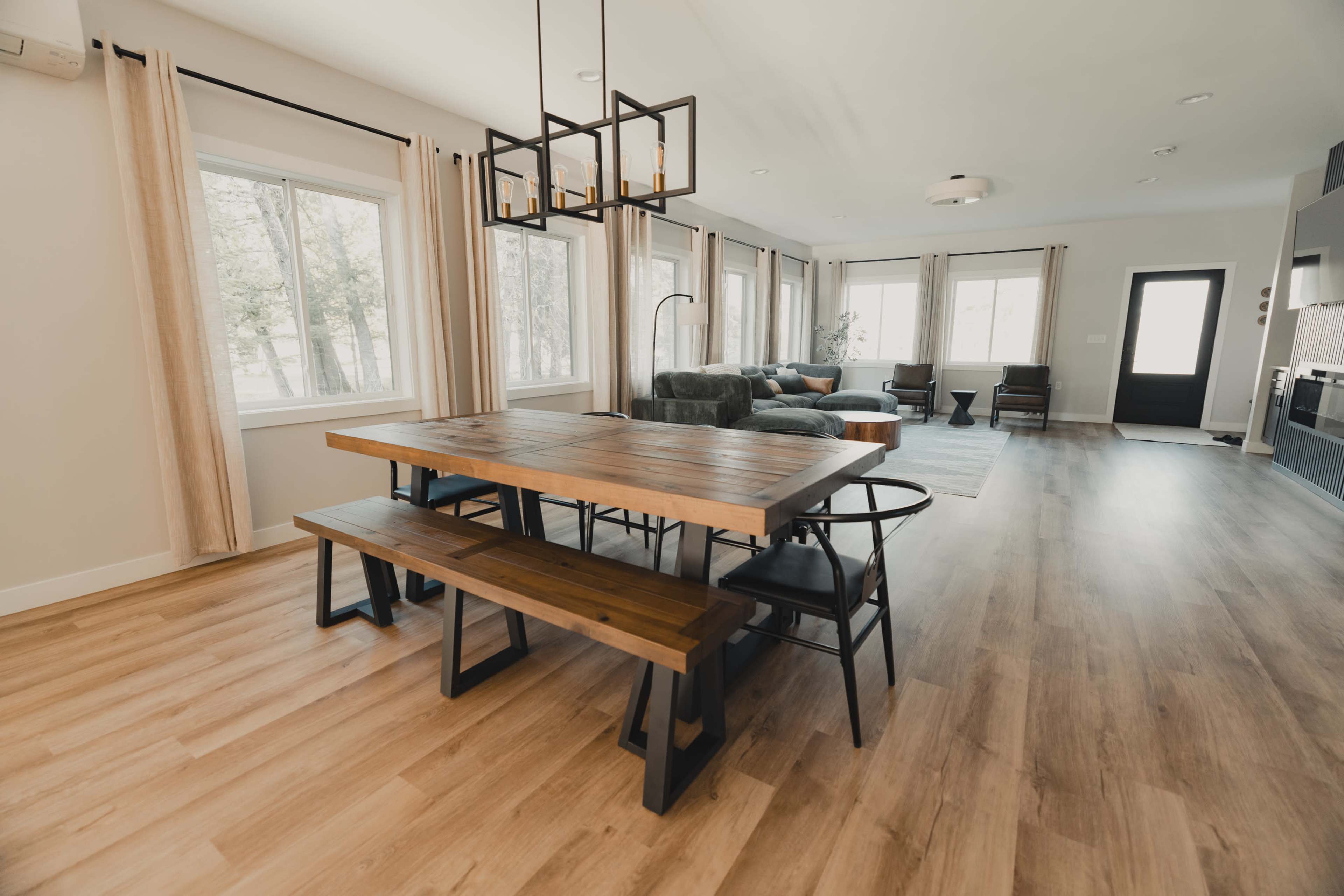 A modern interior features a wooden dining table with benches, surrounded by large windows and a spacious living area with a sofa and coffee table.