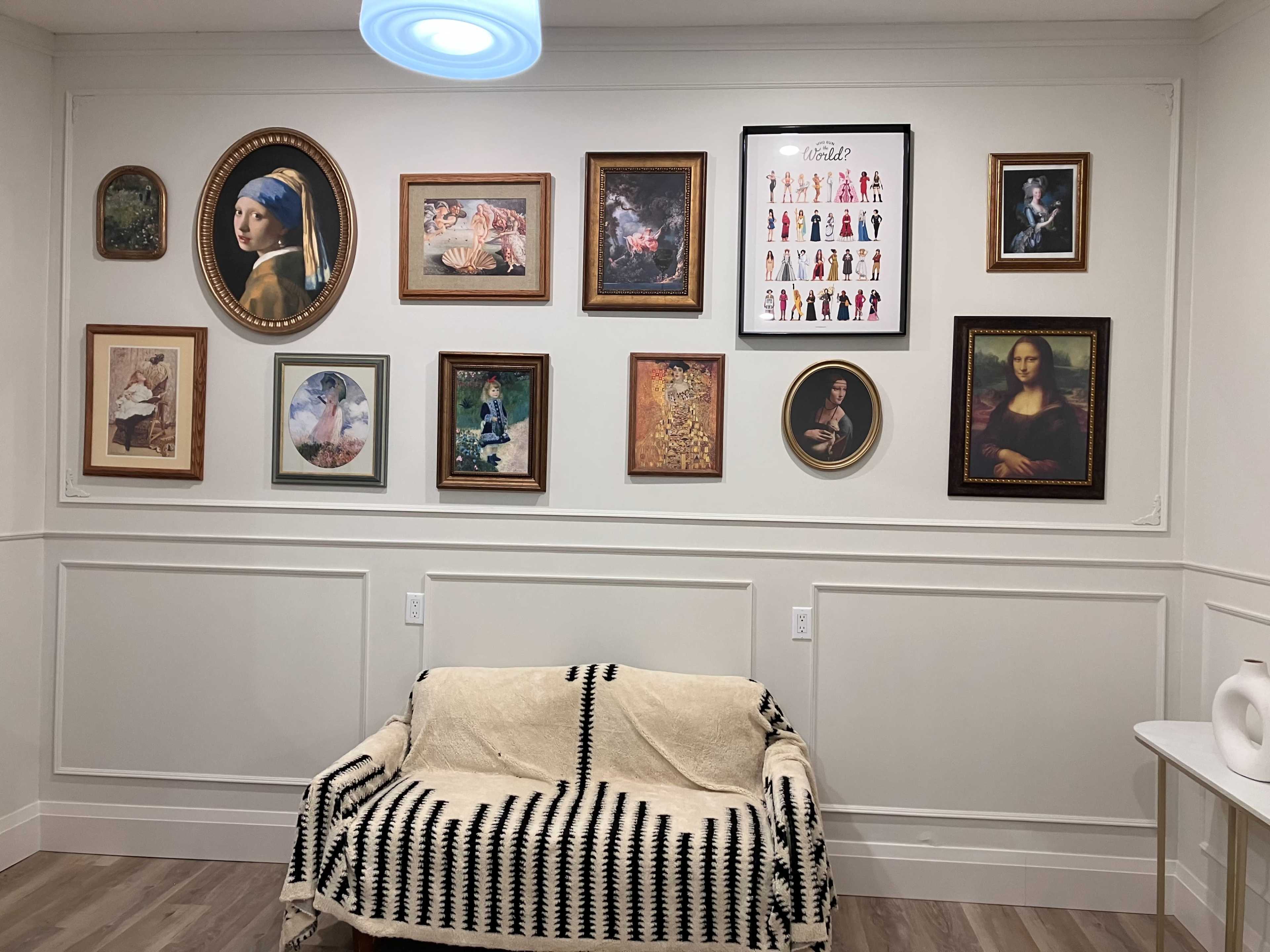 The image shows a wall adorned with various framed artworks, including classical paintings and colorful illustrations, above a simple couch covered with a patterned throw.