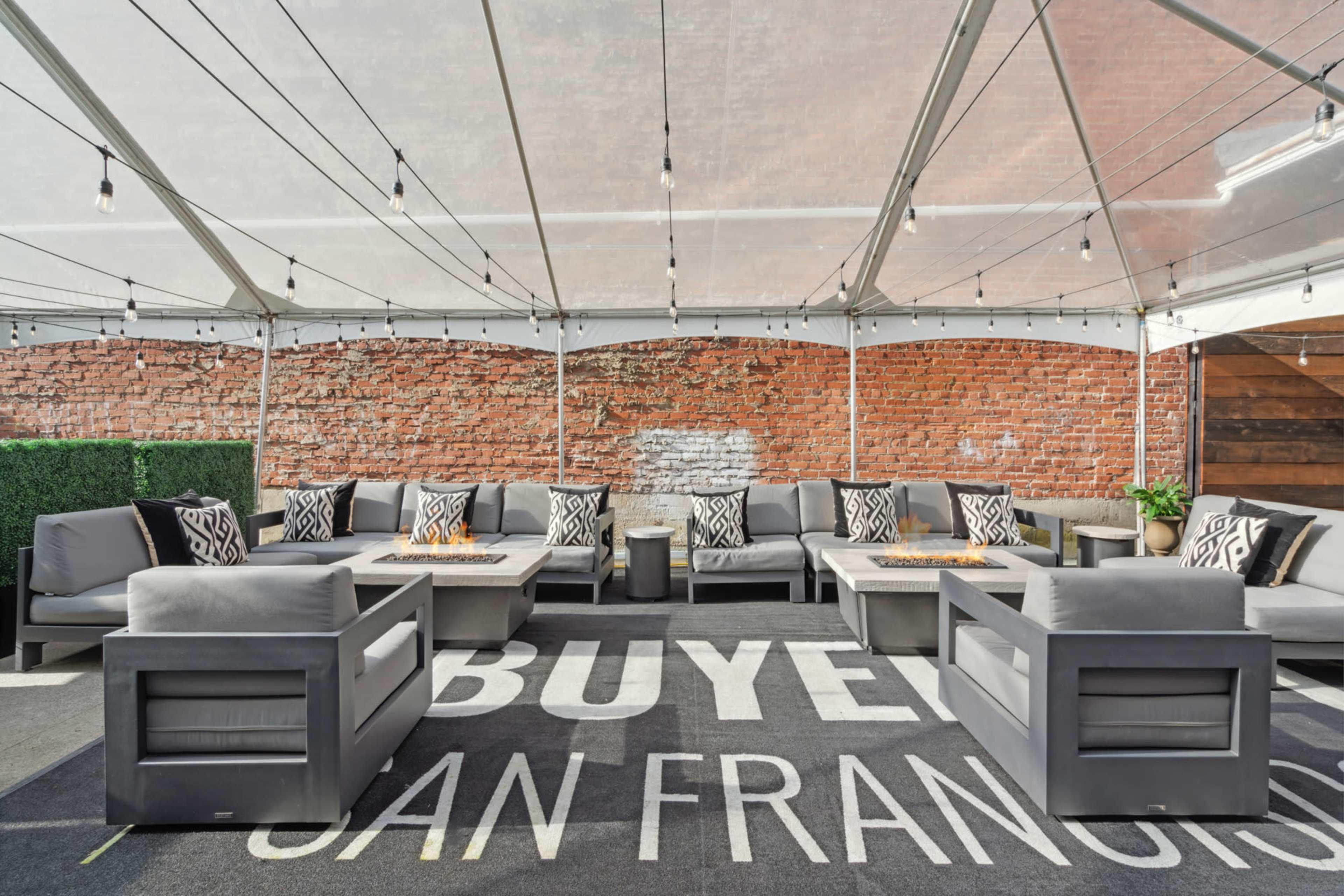 A spacious outdoor lounge area under a tent features gray seating arrangements with patterned cushions and a brick wall backdrop.