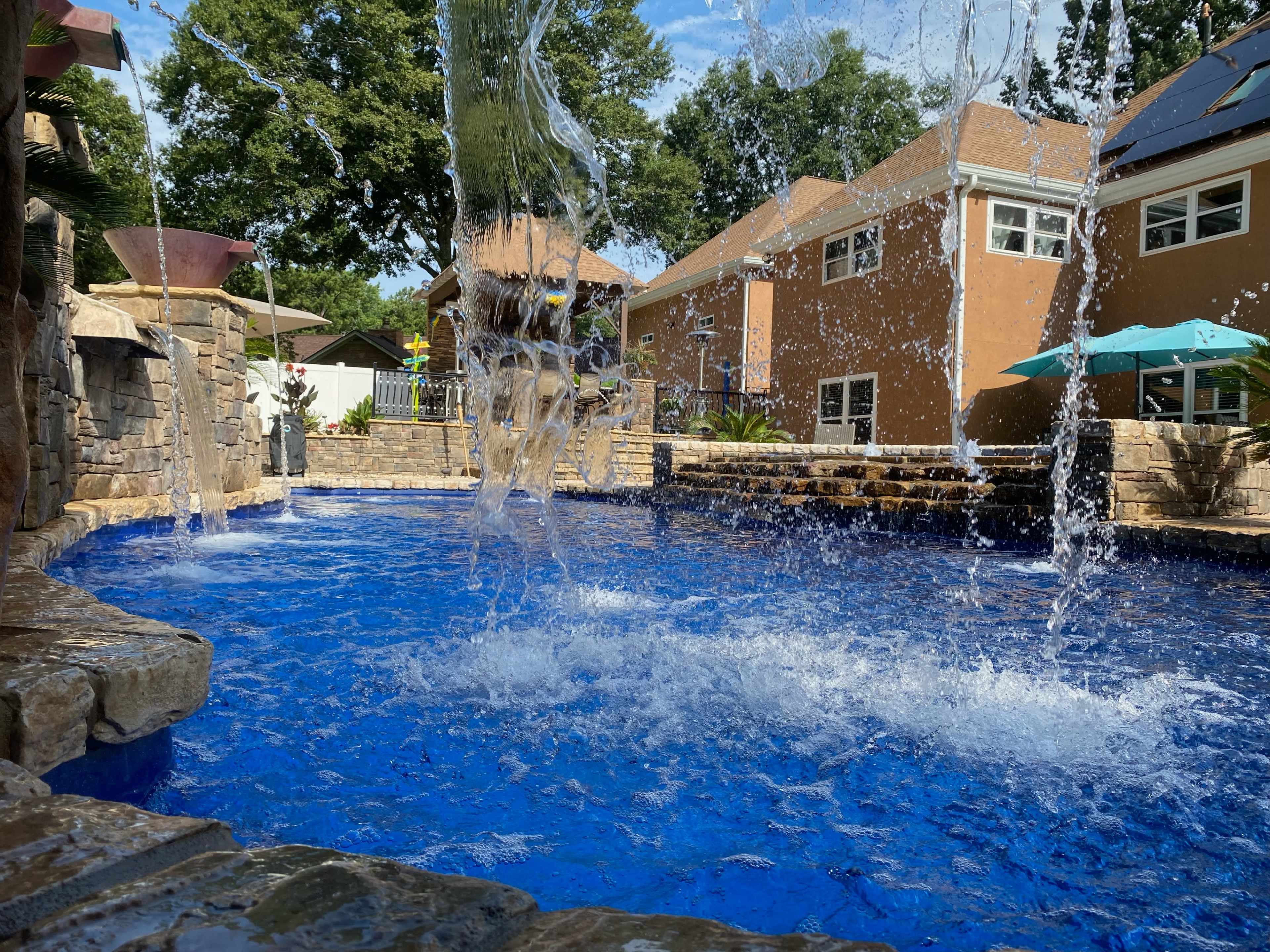 A swimming pool with a stone waterfall features bright blue water and surrounded by landscaped areas and residential buildings.