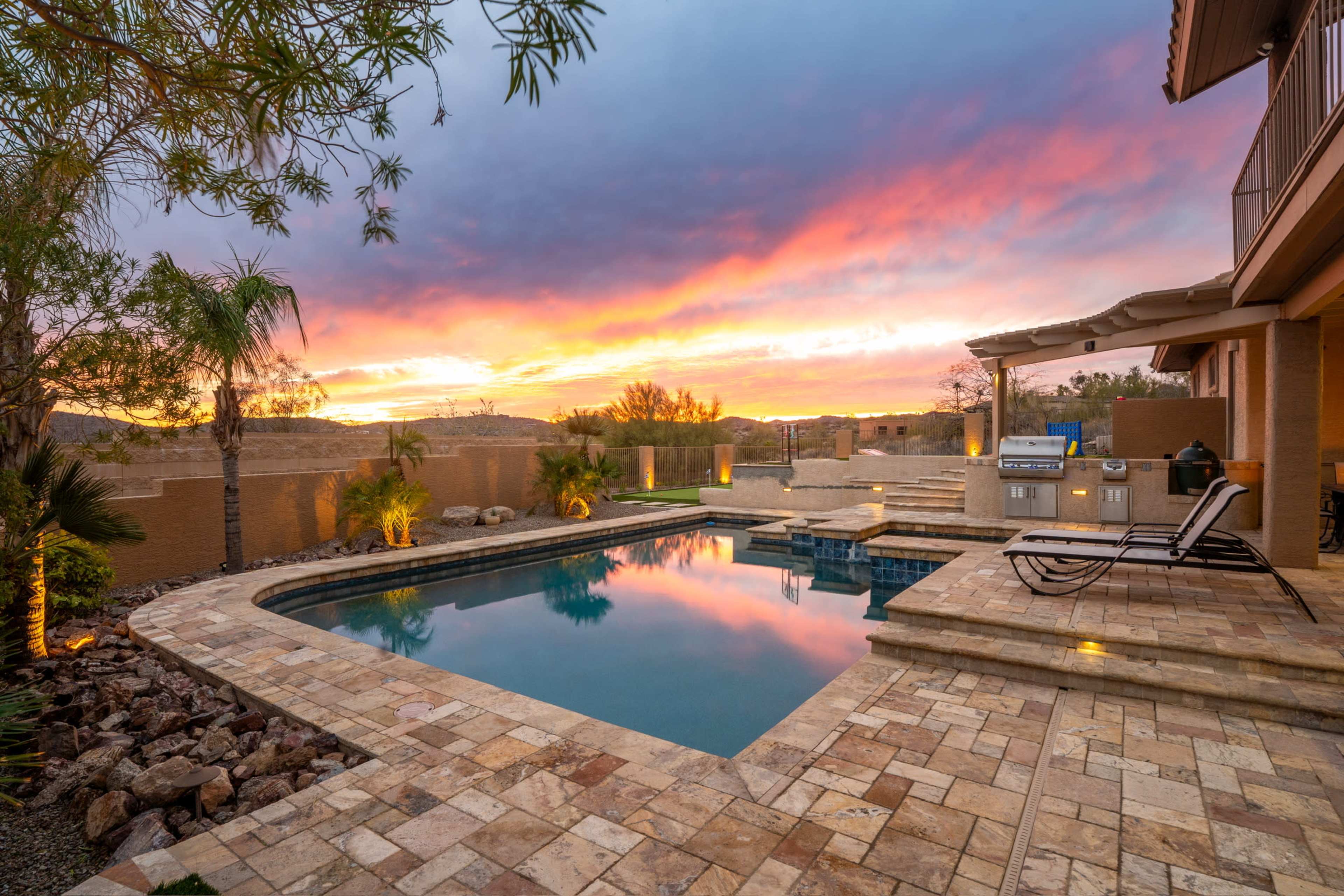 The image shows a backyard pool area with stone tiles and lounge chairs, reflecting a vibrant sunset in the sky above.