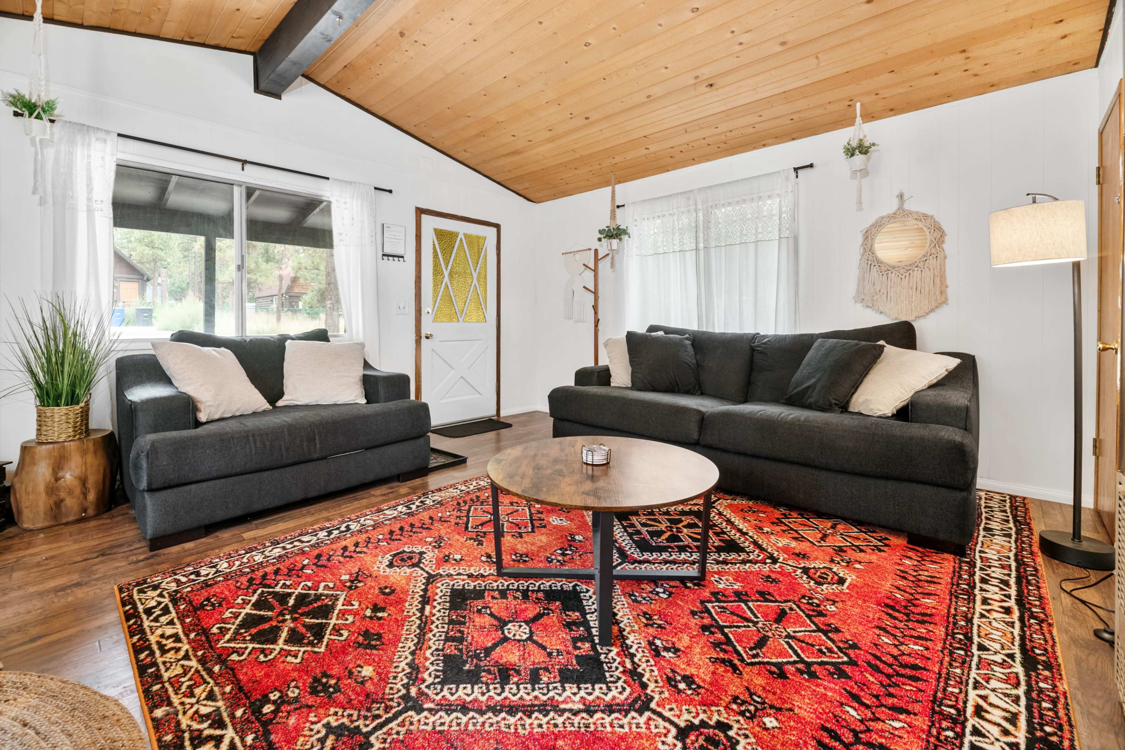 The living room features two dark sofas, a round coffee table, and a patterned rug under a wooden ceiling with large windows and a front door visible.