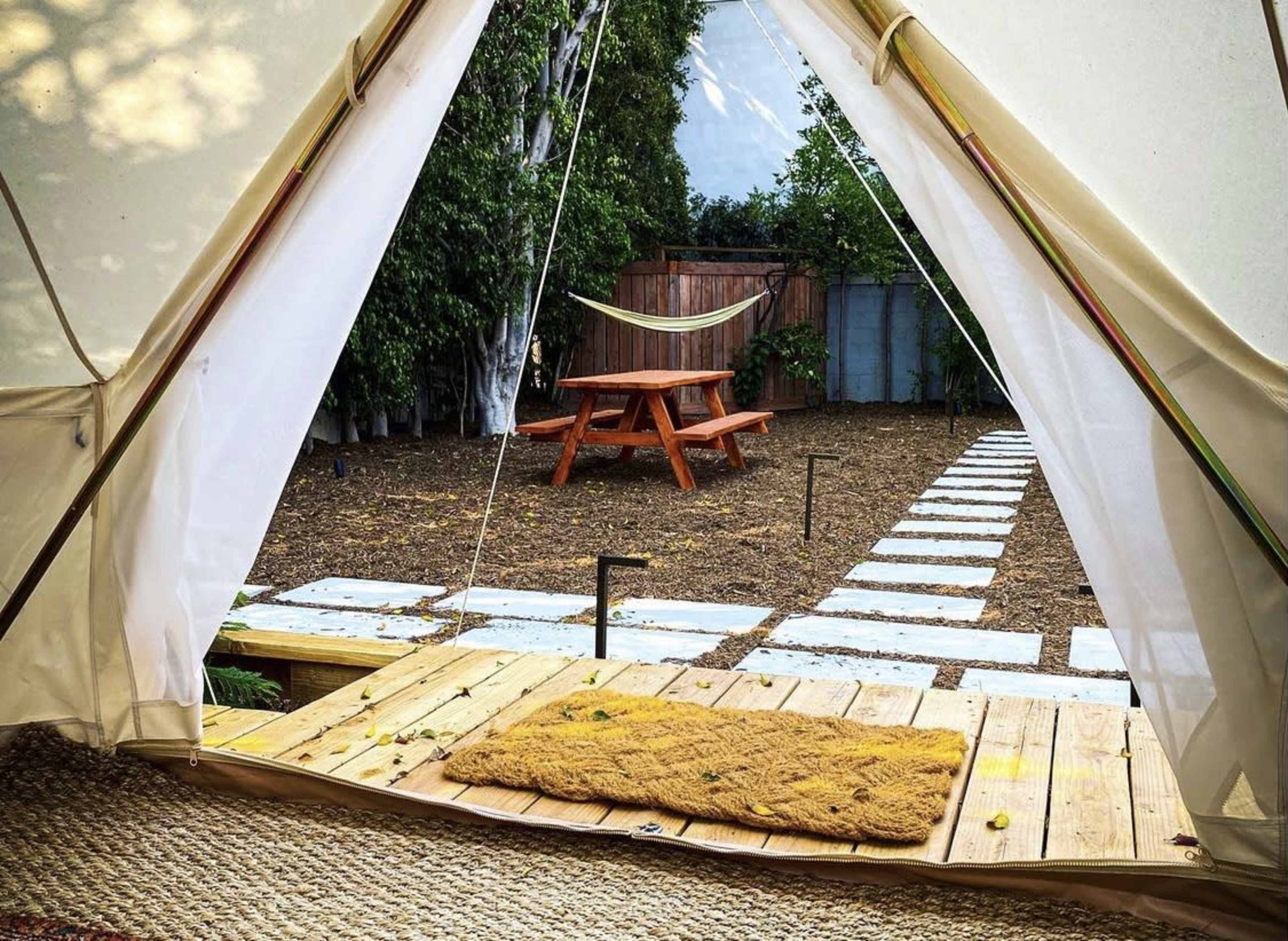 A view from inside a tent showing a wooden deck, a welcome mat, a stone pathway, a picnic table, and a hammock in a well-maintained yard.