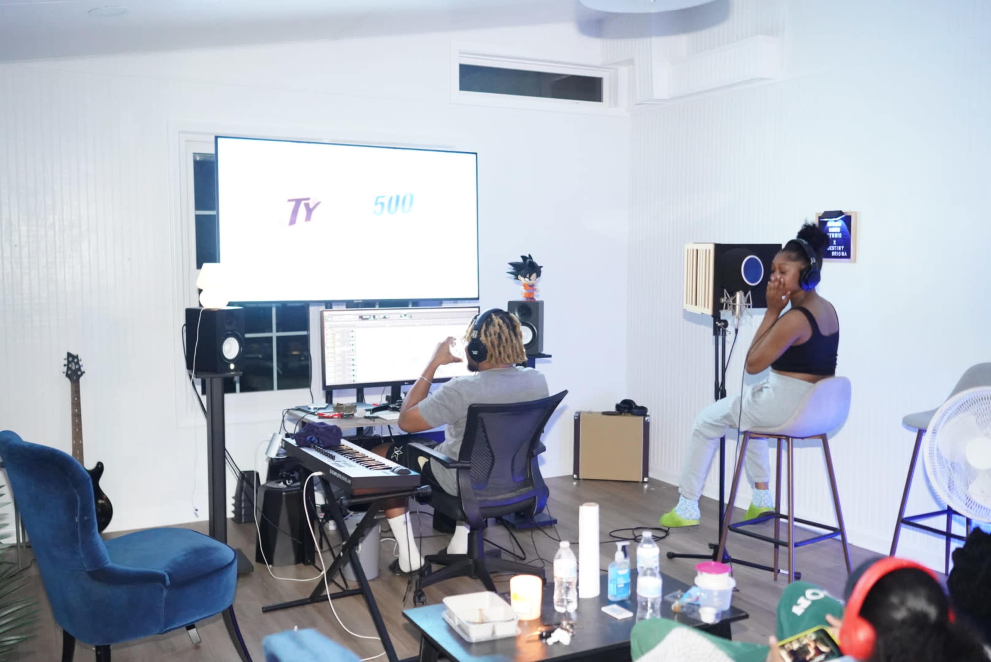 A person with curly hair sits at a computer in a music studio while another person observes from a barstool.
