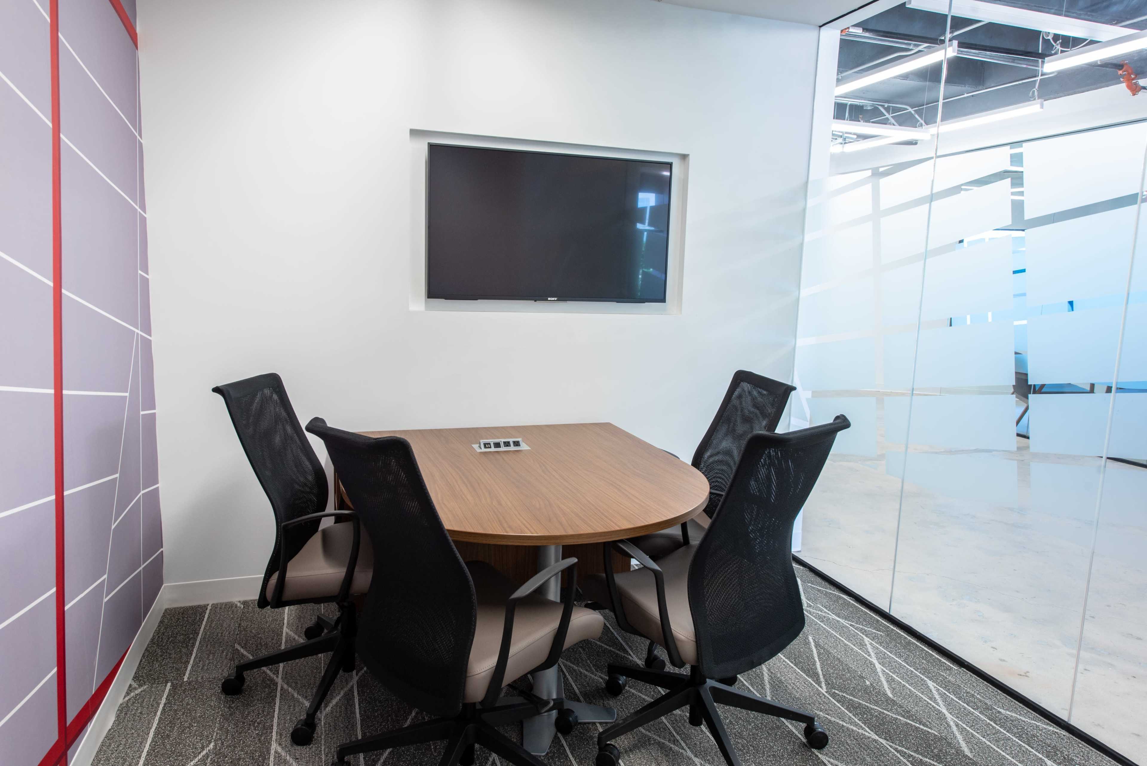 A small conference room features a round table surrounded by four chairs, with a wall-mounted television and a glass partition in the background.