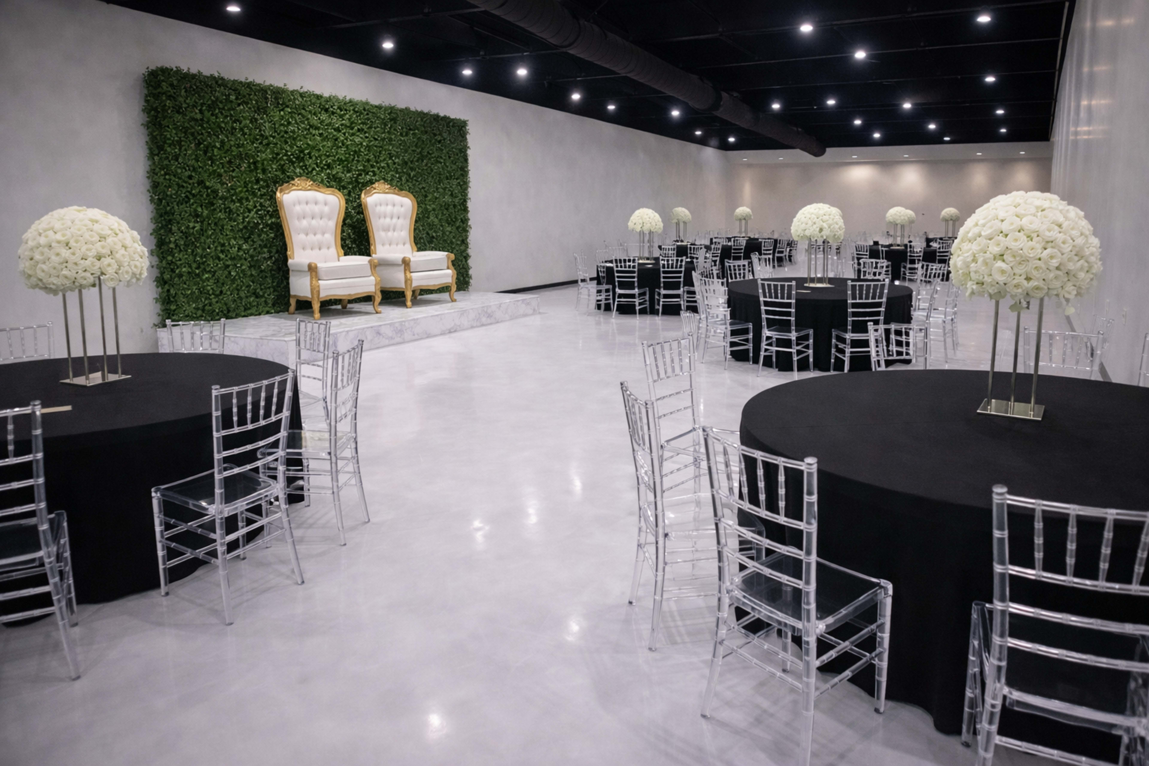 A spacious event room features round black tables with transparent chairs, and a decorative backdrop of greenery behind two ornate golden thrones.