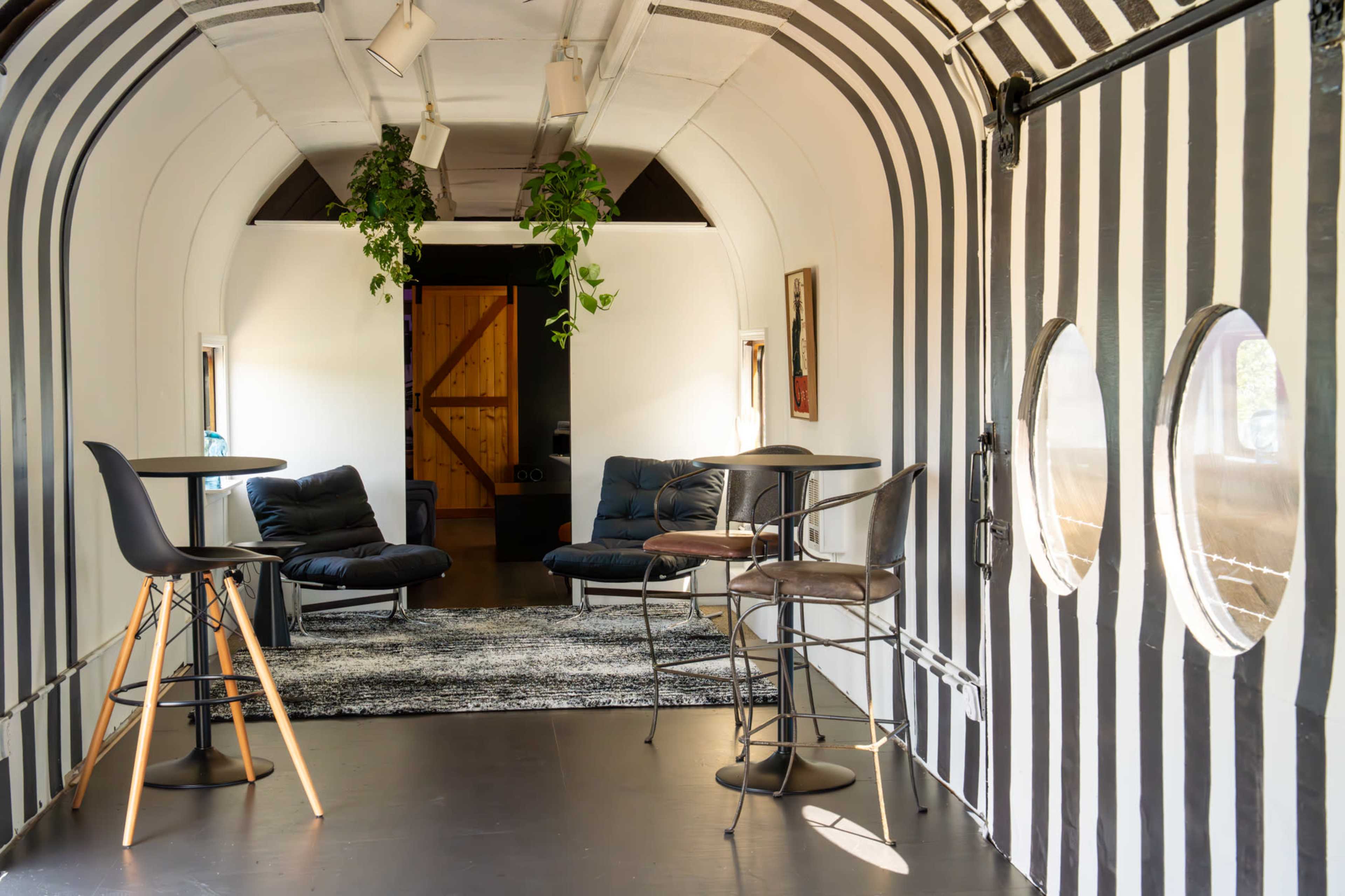 The image shows a room with striped walls, featuring two tall tables and chairs, a seating area with black sofas, and a doorway leading to another space.