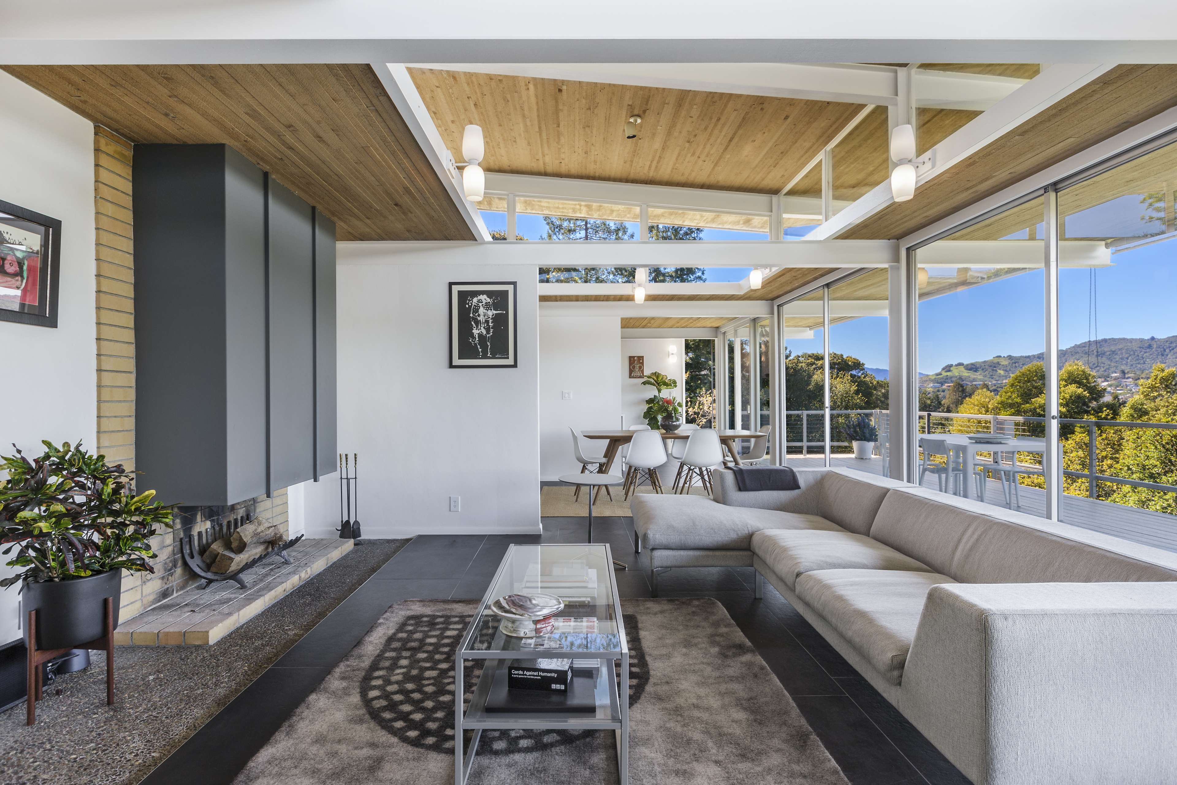 A modern living room featuring large windows, a gray sofa, a coffee table, and a fireplace, with a view of greenery outside.