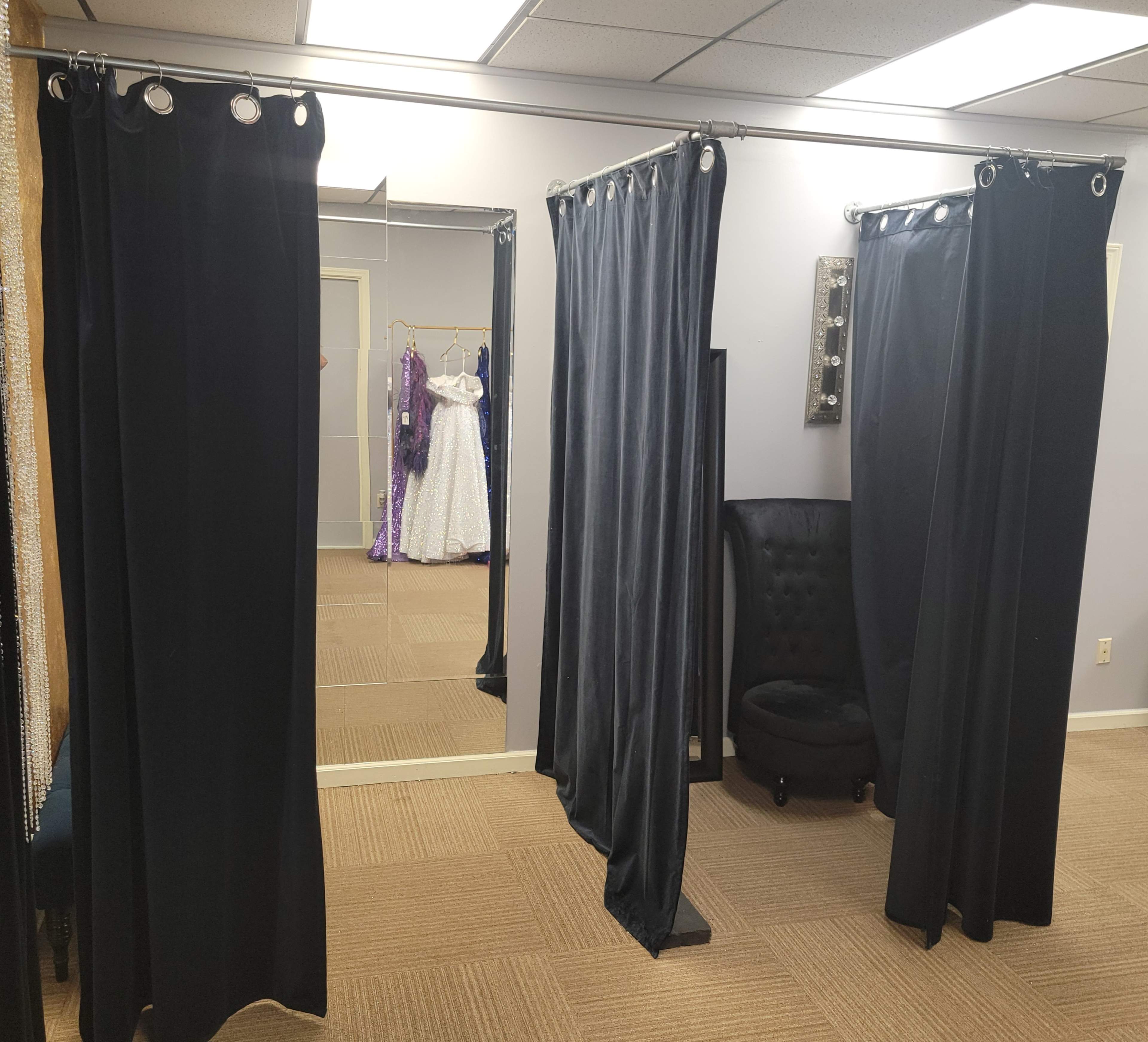 A fitting room area with curtains, a mirror, and a chair, featuring a gown visible in the background.