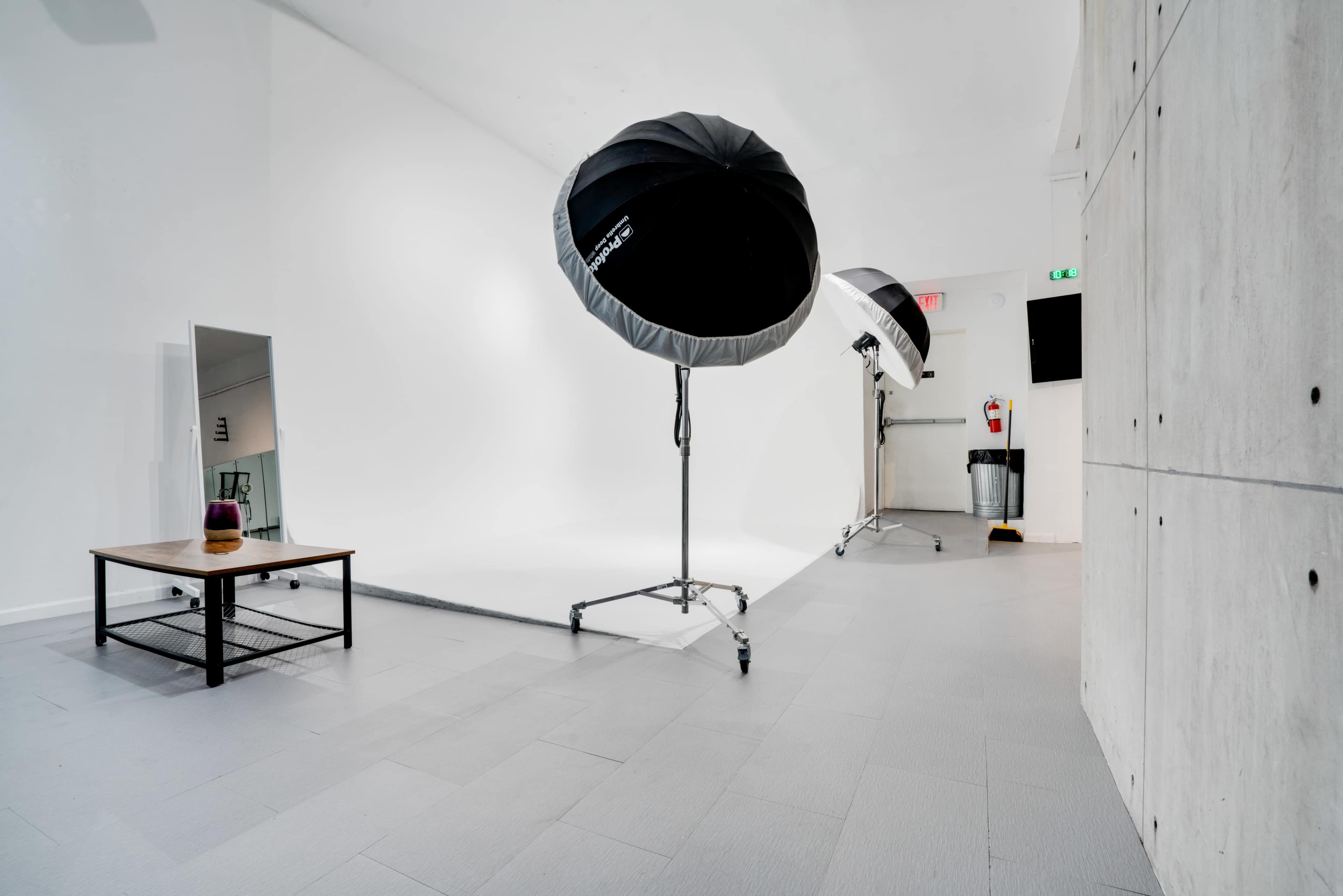 A photography studio featuring softbox lights, a mirror, a table, and a seamless white backdrop.